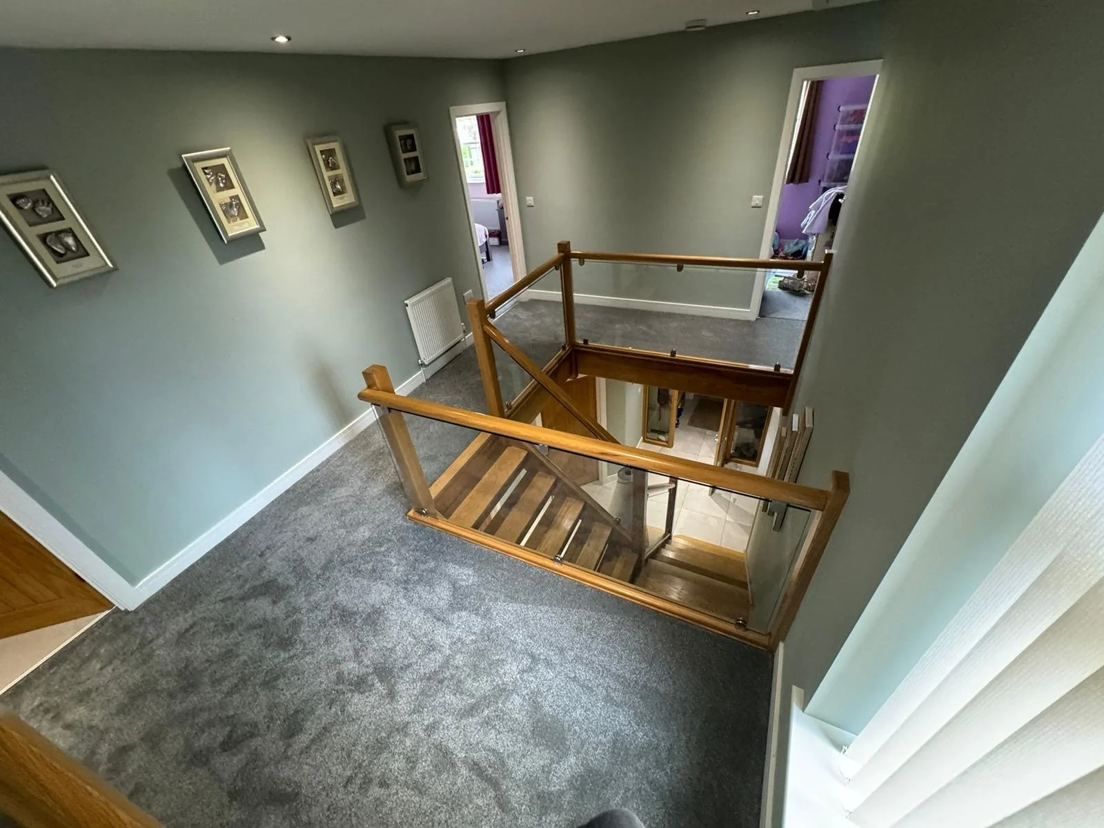 View of a two-story hallway with carpeted floor, green walls, framed photographs, and a wooden staircase with glass panels leading to the lower level, seen from an upper loft area.