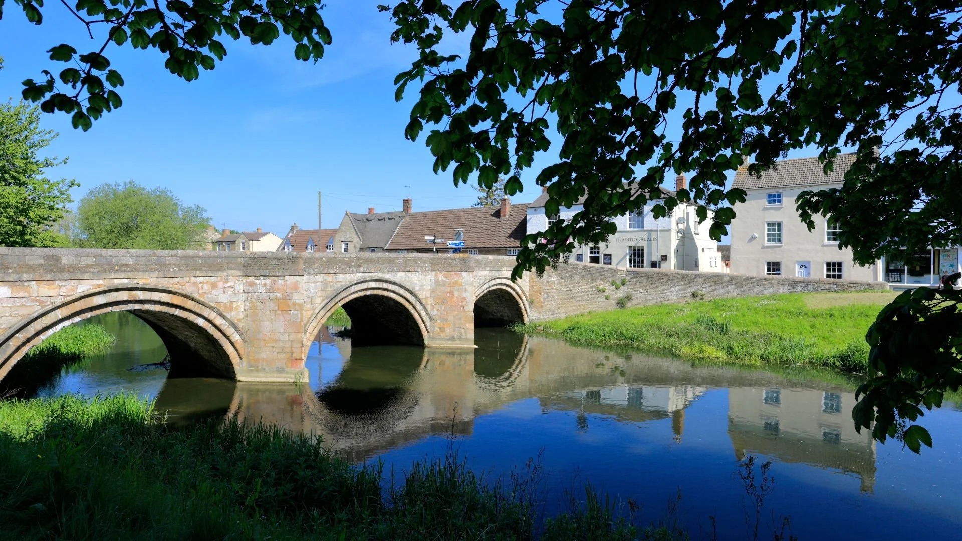 A stone bridge with three arches crossing a calm river, surrounded by green grass and trees, with buildings in the background and a clear blue sky.