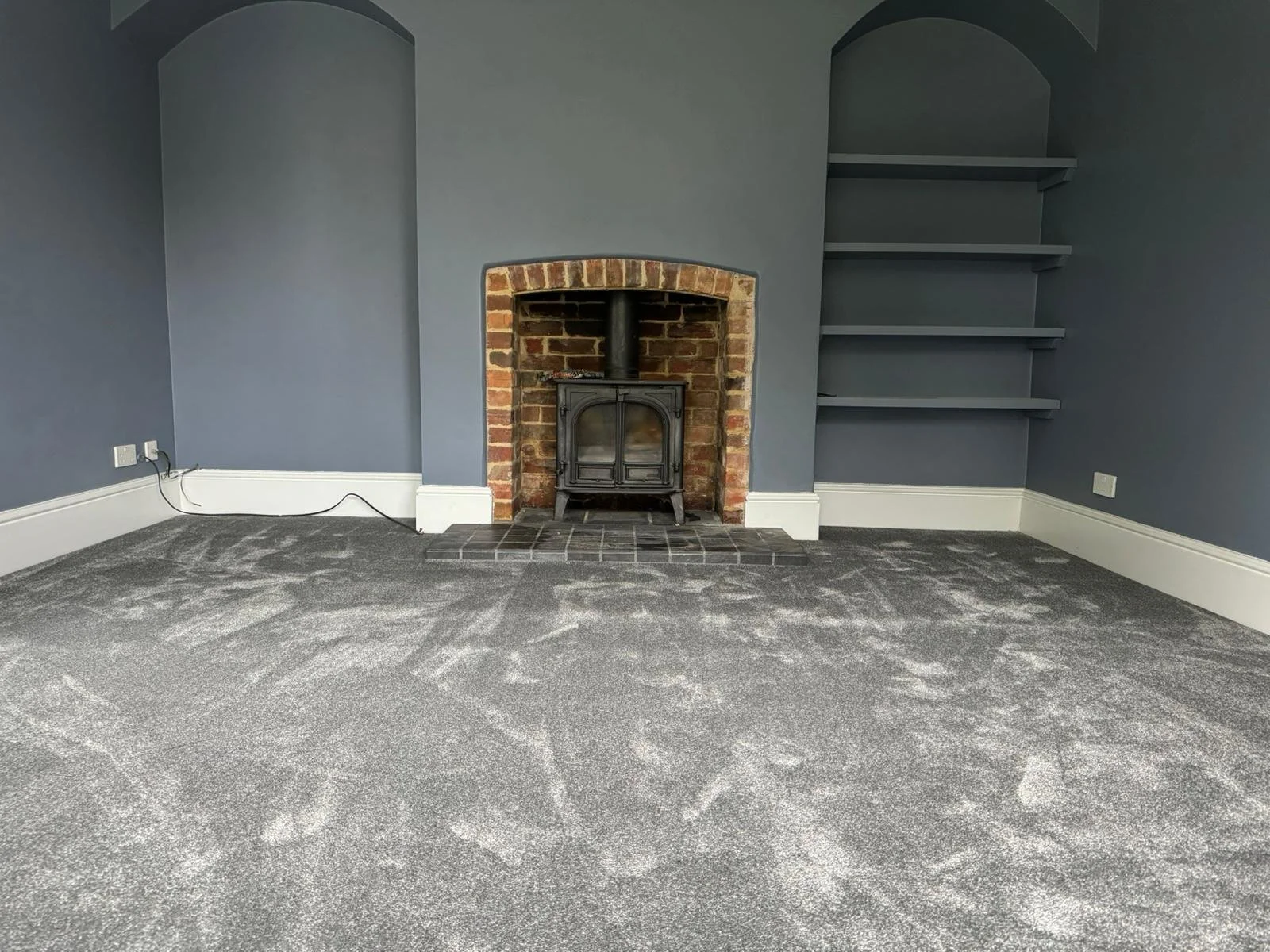 Empty living room with gray walls, a brick fireplace, built-in shelves, and gray carpet flooring.