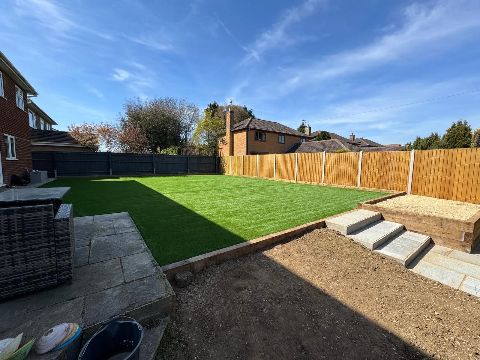 Backyard with freshly laid green artificial grass, bordered by wooden fences, a patio area with stone steps, and neighboring houses under a blue sky.