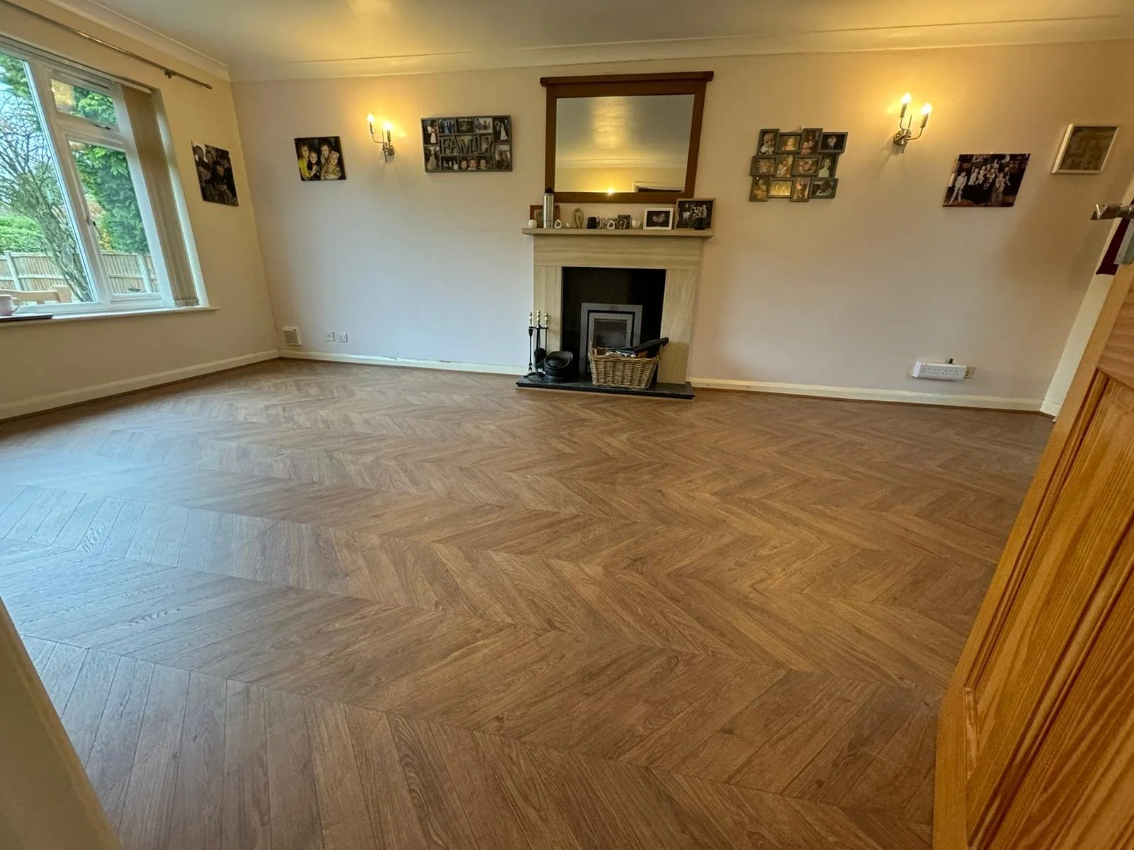 Empty living room with wooden herringbone patterned floor, cream-colored walls, a fireplace with a basket and fireplace tools, family photos and picture frames on the wall, a window to the left with a view of trees and a fence outside, and wall-mount