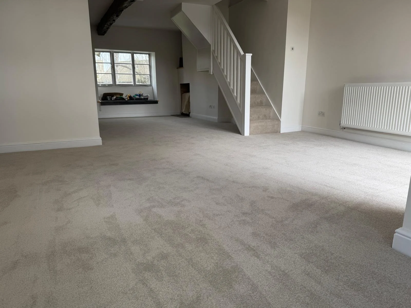 Empty room with beige carpet, white walls, a staircase with beige carpet, radiator, window, and some clutter on a black shelf.