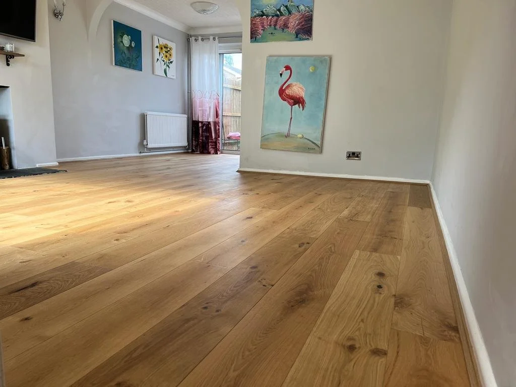 A living room with hardwood floor, white walls, colorful floral artwork, a door with sheer curtains leading outside, and a pink flamingo painting on the wall.