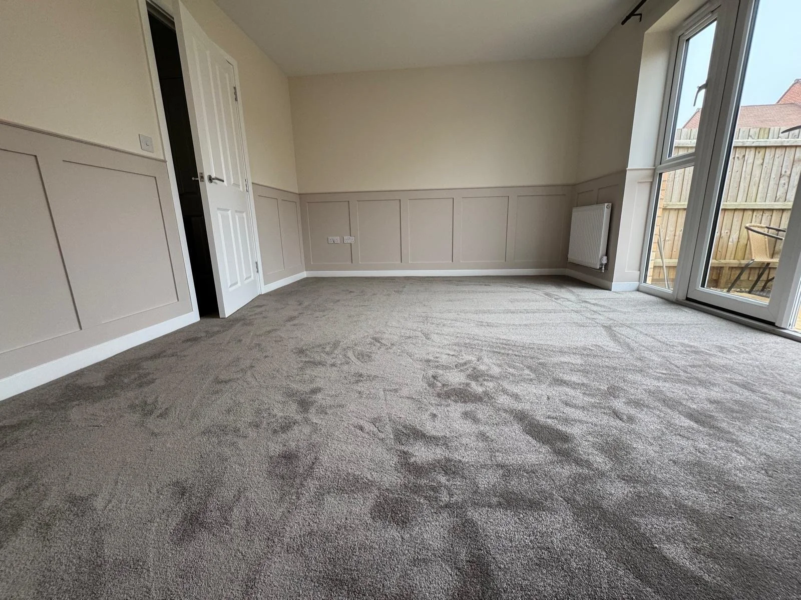 Empty living room with beige carpet, cream-colored walls with wall paneling, open door to a closet, large glass sliding door leading to an outdoor patio, and visible outdoor fence and furniture.