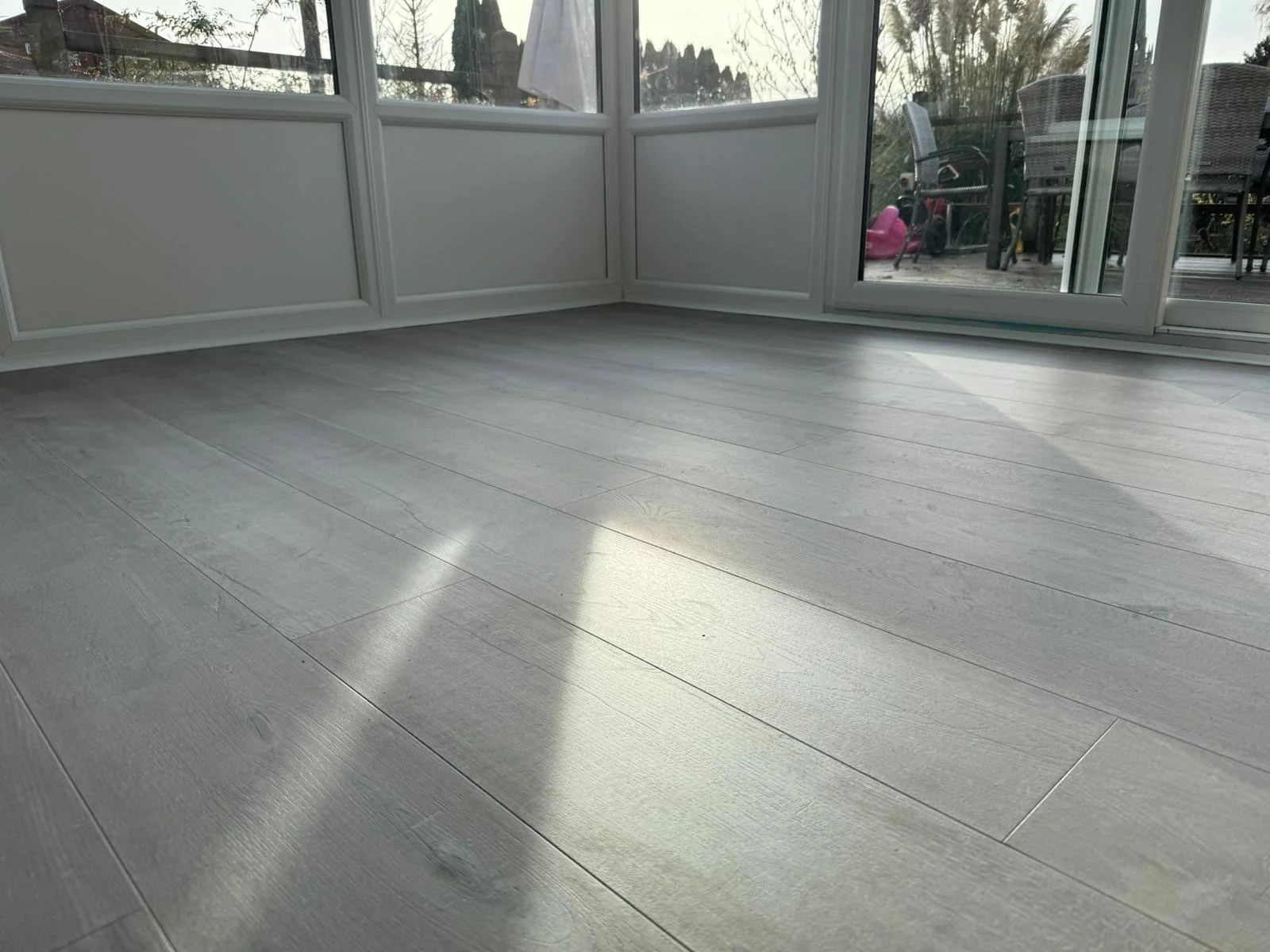 Flooring inside a sunroom or enclosed porch with light-colored wood or laminate planks and large windows showing outdoor patio furniture and play equipment.