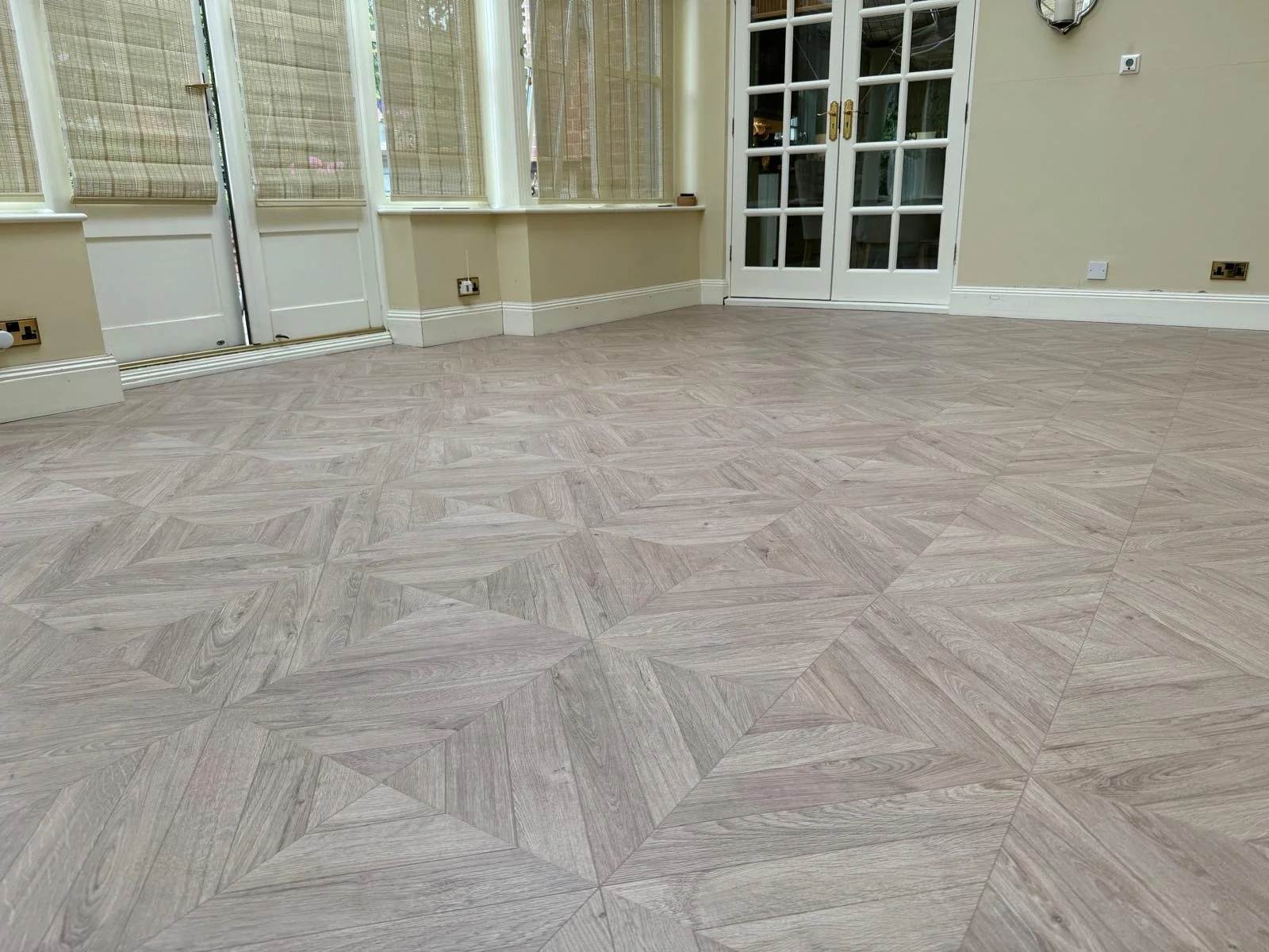 Empty room with light wood patterned flooring, cream walls, and double glass doors leading to an outdoor area, with some electrical outlets and switches visible.