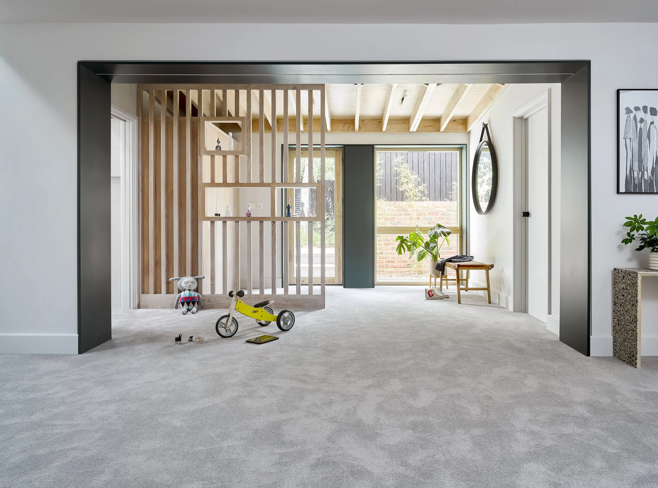 Spacious, modern living room with light-colored walls and gray carpet. Features a wooden rail partition, a sliding door, a large window, and minimal furniture including a small bench with a blanket, a potted plant, and a square side table. Toys, including a stuffed animal, a balance bike, and a tablet, are scattered on the floor.