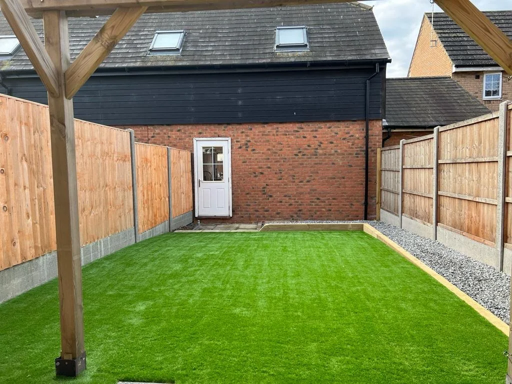View of a backyard with a freshly laid green artificial grass lawn, surrounded by wooden fencing, with a brick house wall and a door in the background, and a partially visible wooden structure framing the top of the image.