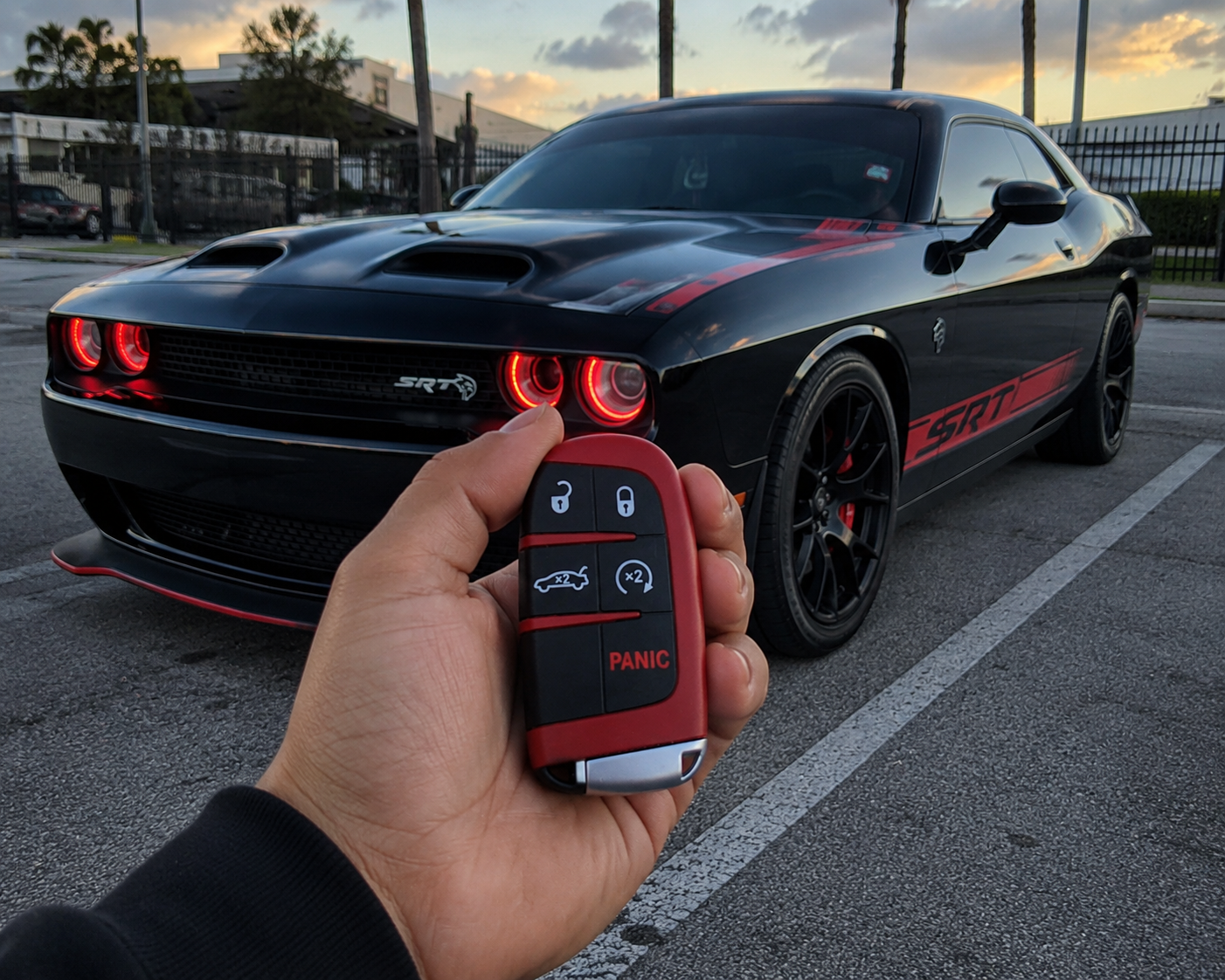 A black Dodge Challenger SRT muscle car with red accents, parked in a lot during sunset. A person's hand holds a red key fob with control buttons in front of the car.