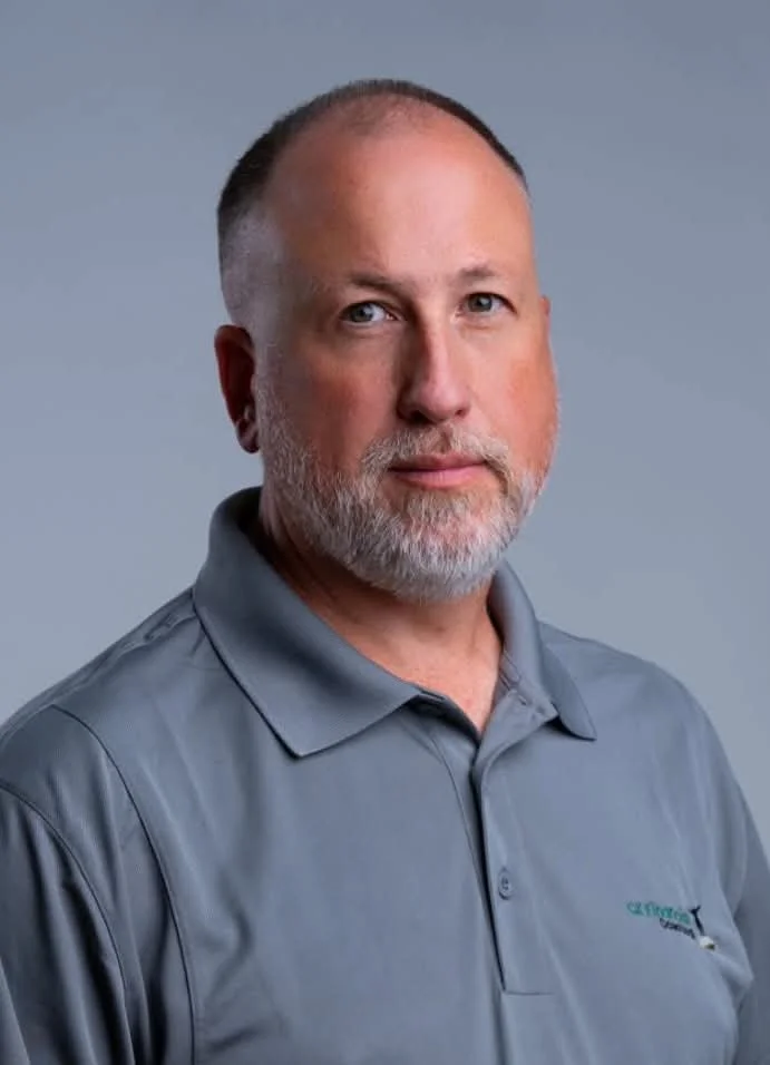 A middle-aged man with a grey beard and short hair, wearing a grey polo shirt with an embroidered logo, posing against a plain, light grey background.