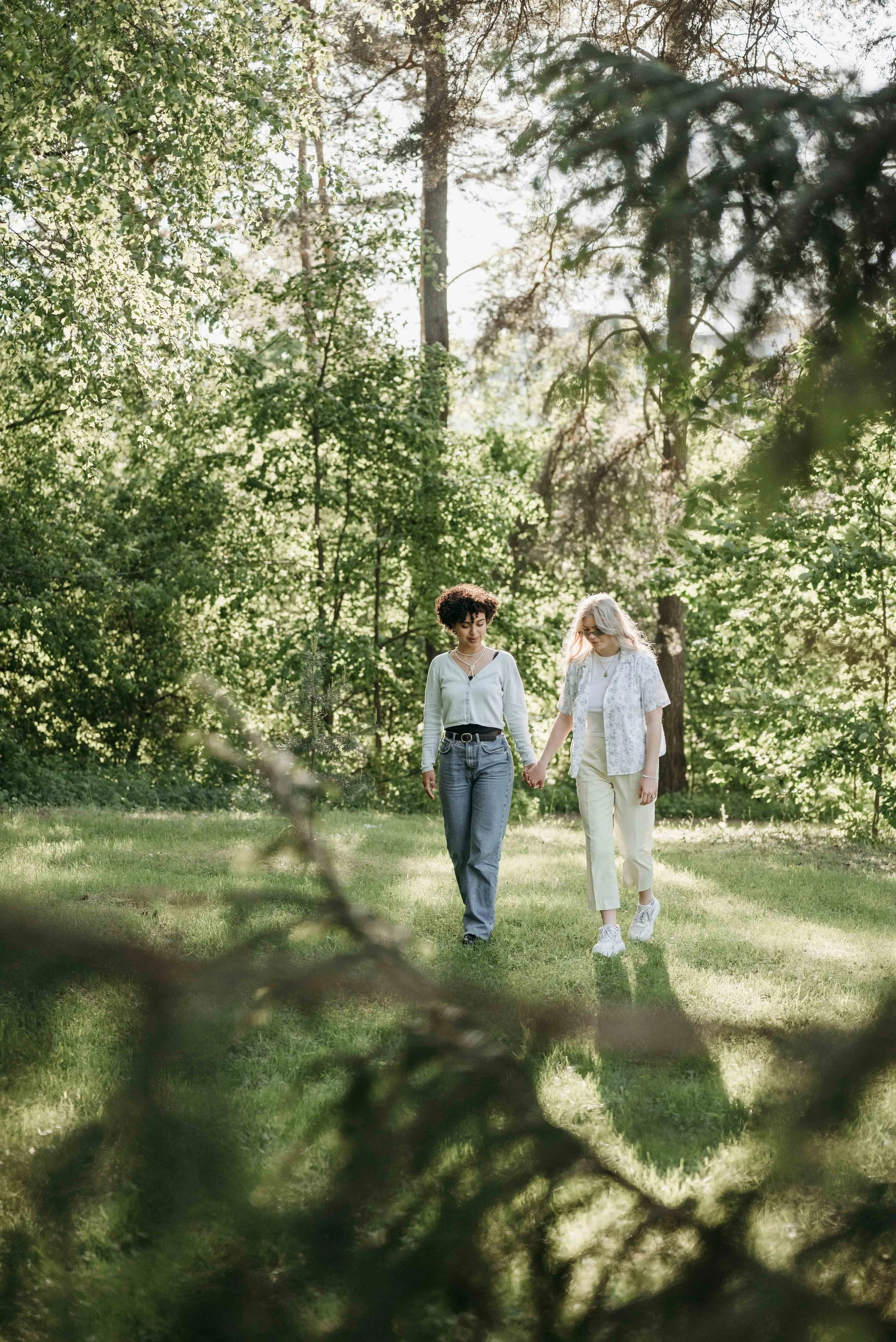 Two women walk hand-in-hand through a sunlit forest clearing, surrounded by green trees and foliage.