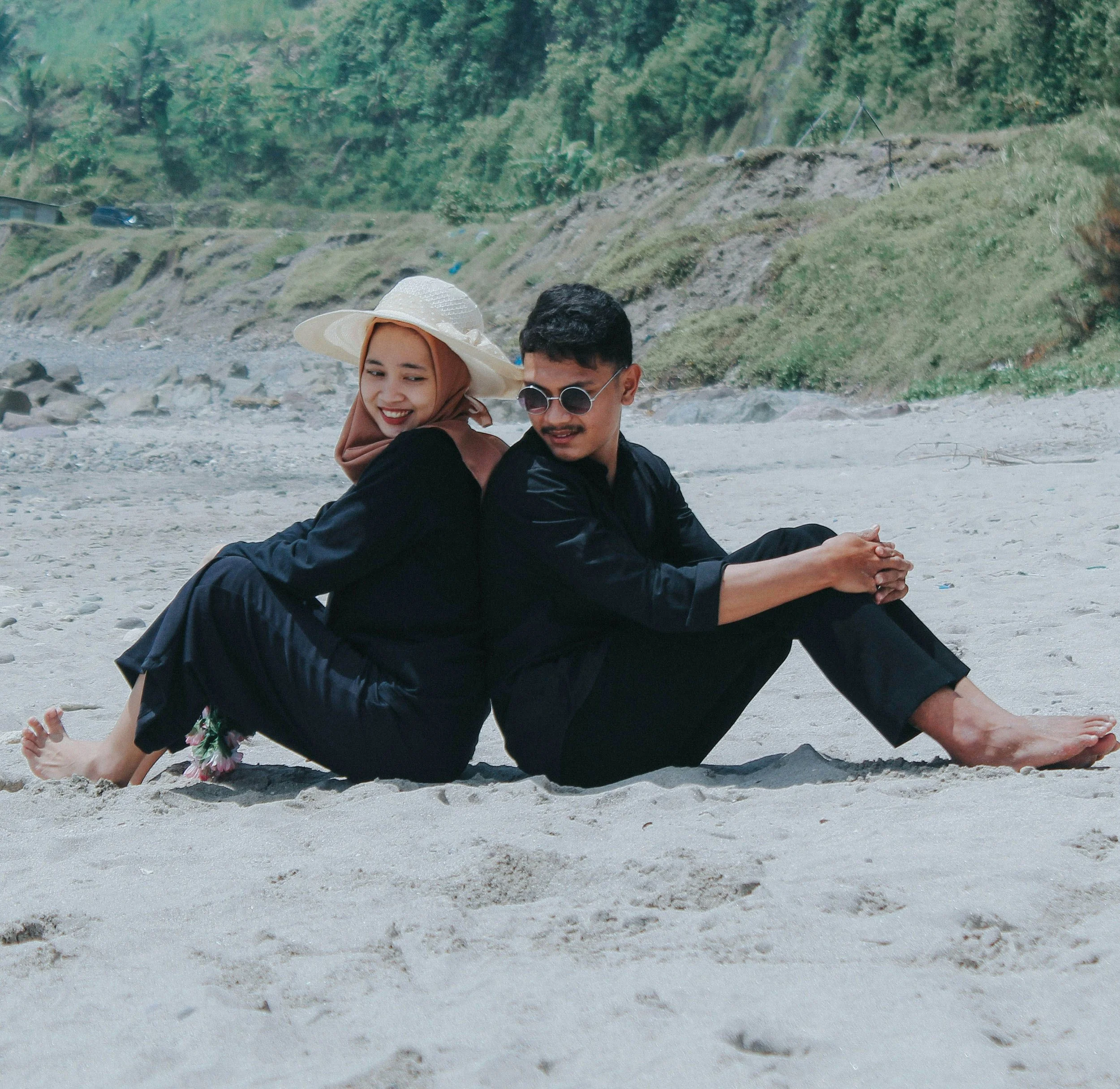 A man and woman are sitting back-to-back on a sandy beach, smiling. The woman wears a wide-brimmed hat and a hijab, while the man wears sunglasses and black clothing. In the background, there are green hills and some rocks.
