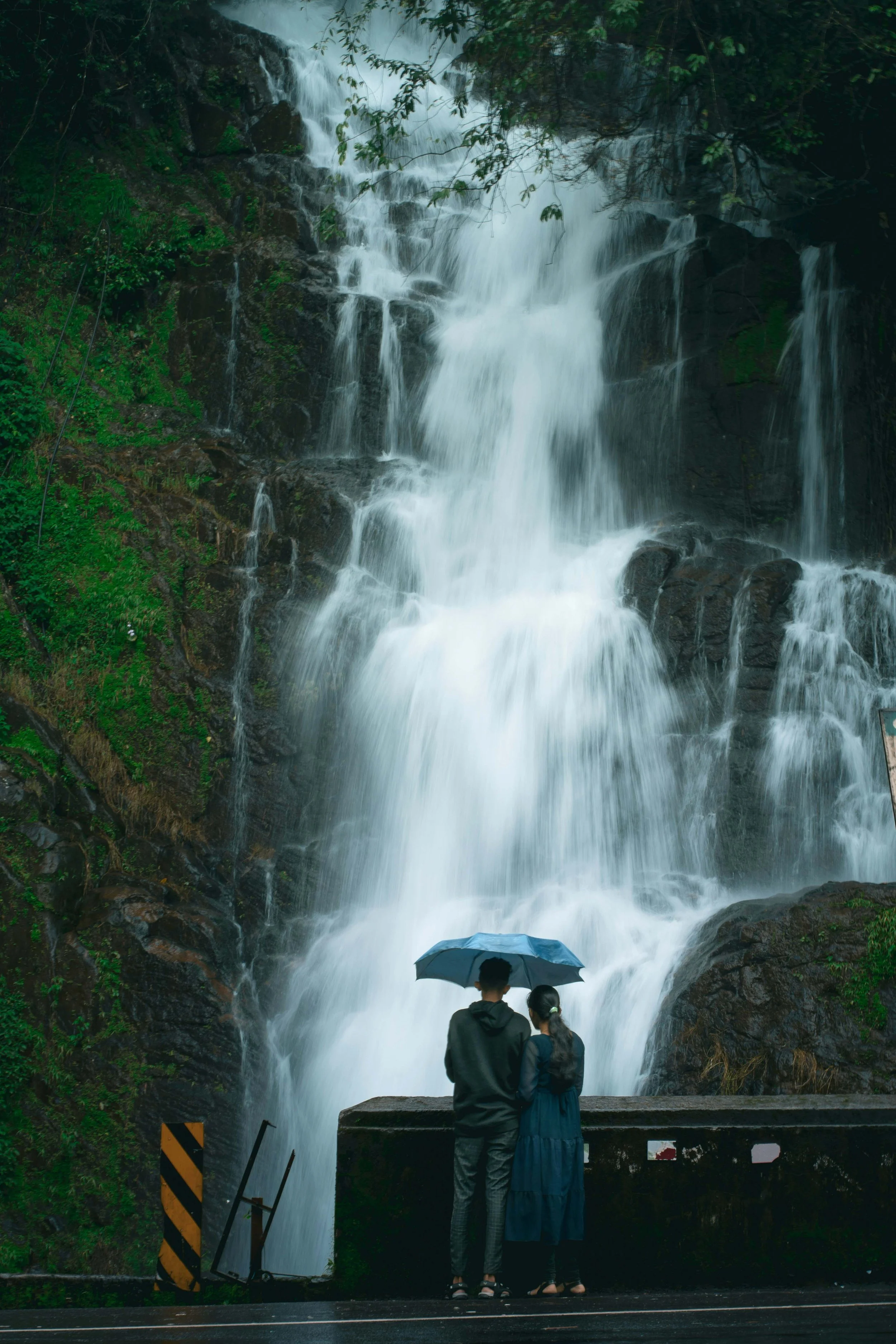 A couple stands on a wet road holding an umbrella, watching a large waterfall with flowing water cascading down rocky cliffs, surrounded by green trees.