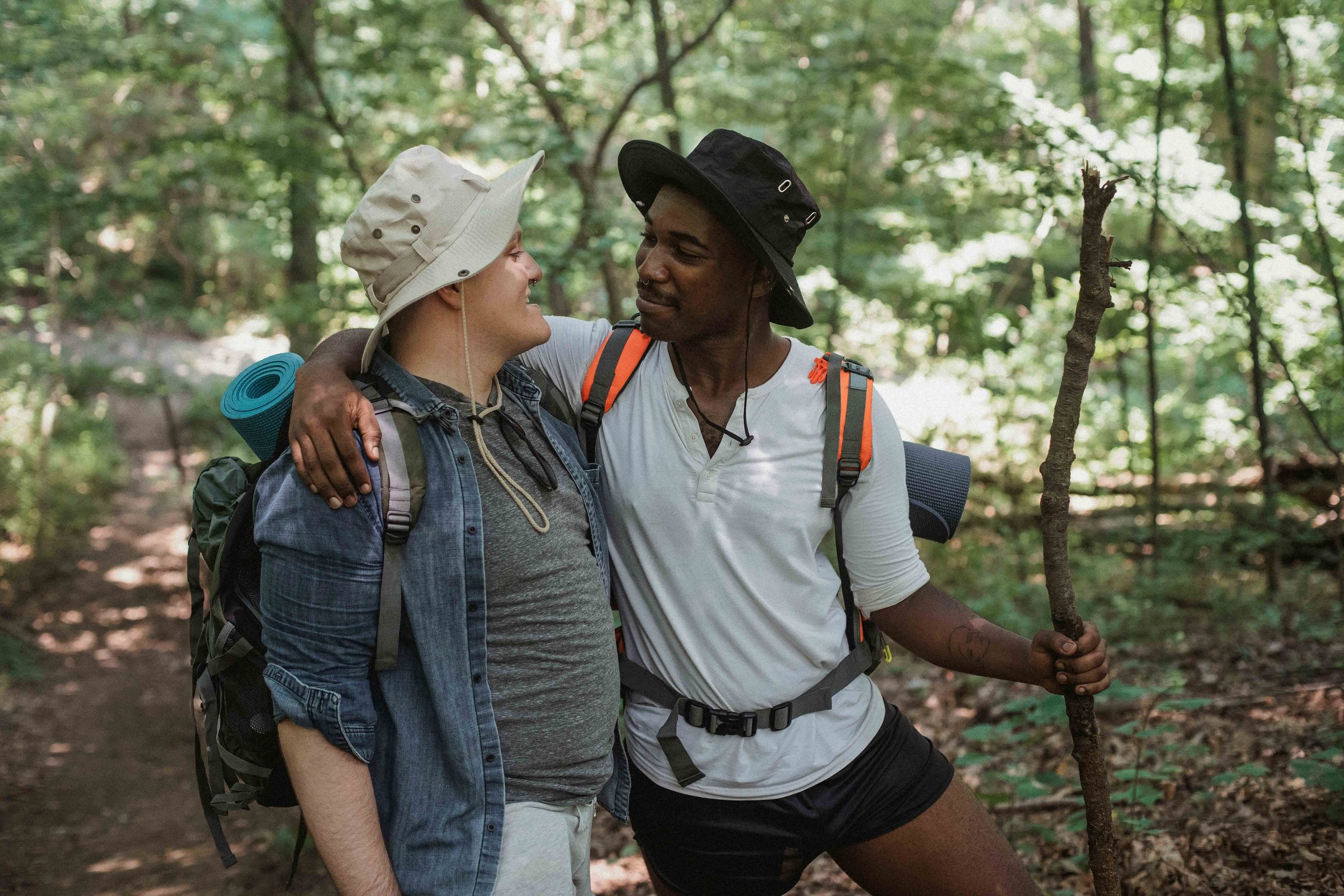 Two hikers in a forest, smiling and looking into each other's eyes, with backpacks and hiking gear, amidst green trees and dappled sunlight.