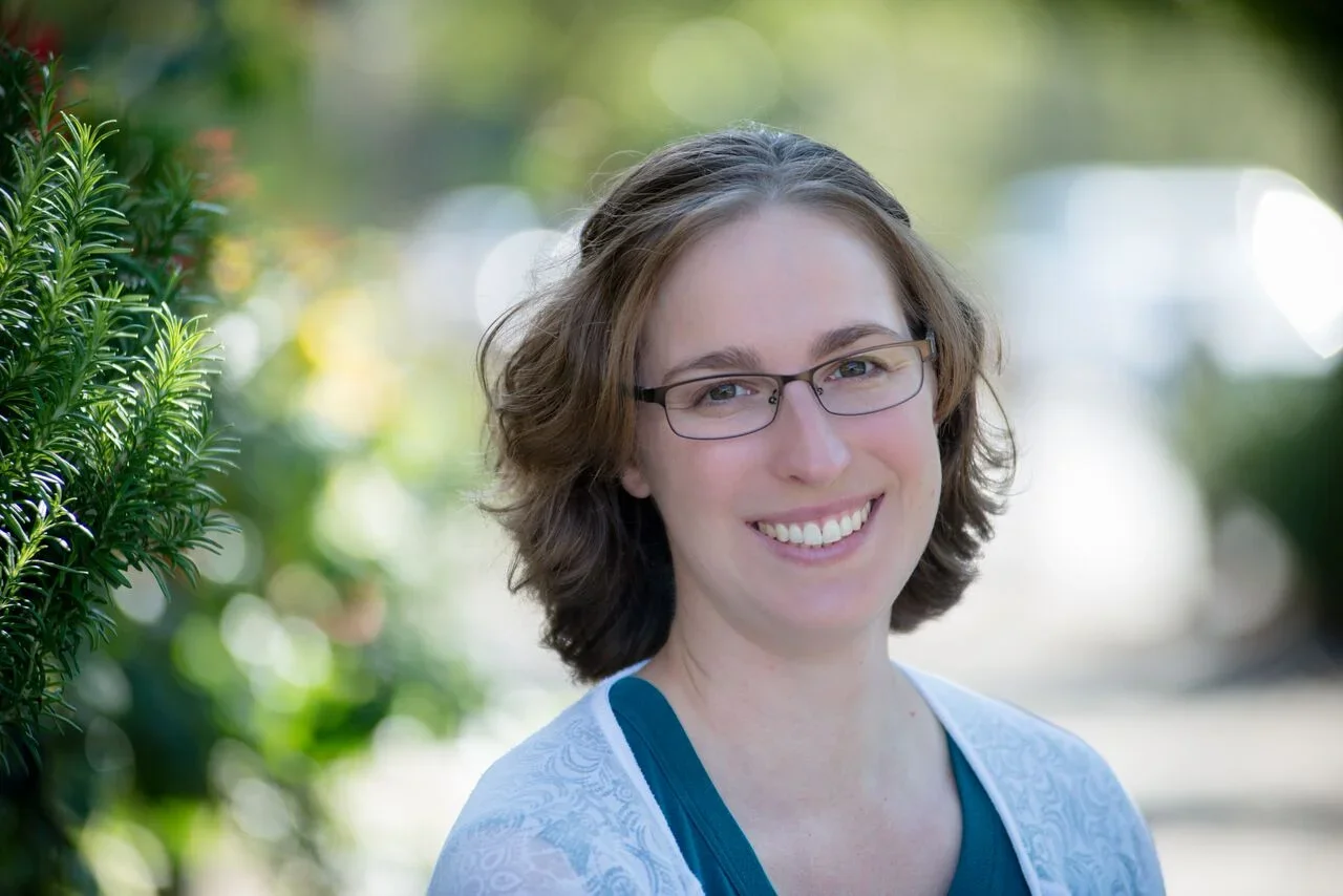 A woman with shoulder-length brown hair, glasses, and a smile, standing outdoors near greenery.