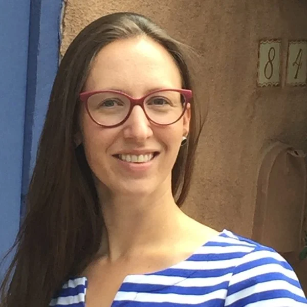 Woman with long brown hair, wearing pink glasses and a blue and white striped shirt, smiling outdoors near a wall with house number signs.