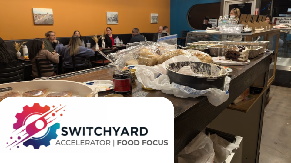 A long wooden table with various plates of food, including cakes and pastries, is in the foreground. In the background, a group of people sit at a table in a colorful room with orange and teal walls, some engaged in conversation.