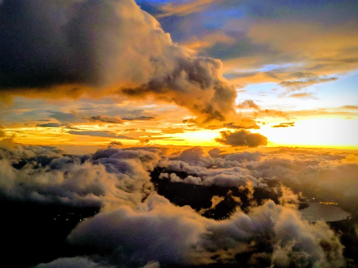 A colorful sky at sunset or sunrise with clouds in shades of dark gray, orange, and yellow, partially illuminated by the sun.