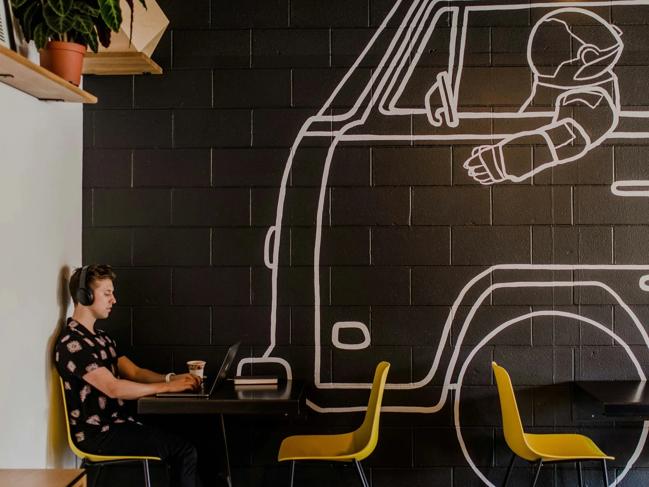 A young man with headphones working on a laptop in a cafe, with a black brick wall featuring a white line art mural of a delivery truck in the background.