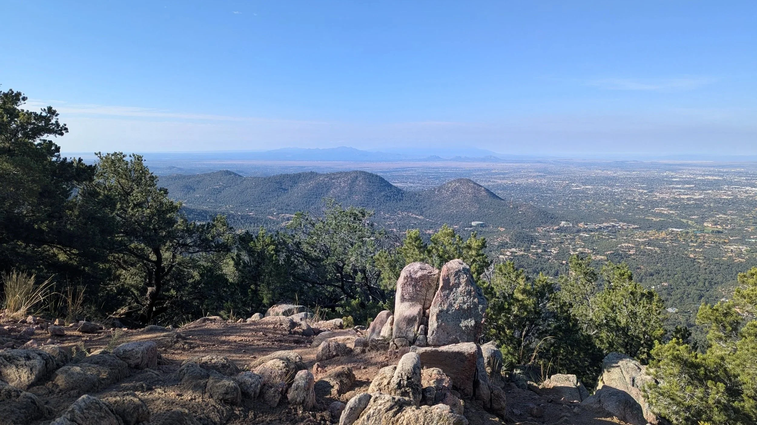Mountain view overlooking a city with trees and rocks in the foreground and a clear blue sky.