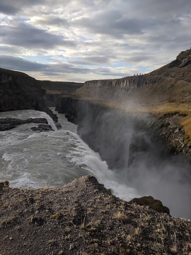 A river flowing rapidly through a canyon with waterfalls, rugged cliffs, and cloudy sky overhead.