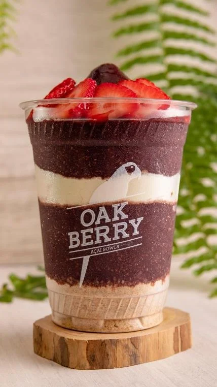 A layered acai bowl with strawberries on top, served in a clear cup with a logo that reads 'OAK BERRY ACAI BOWLS', placed on a wooden coaster, with a fern plant in the background.