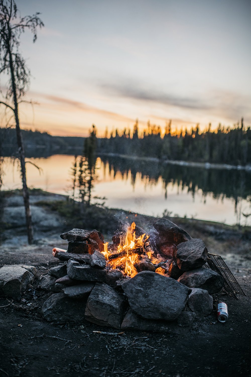 Campfire with flames burning surrounded by rocks near a calm lake at sunset, with trees reflecting in the water.