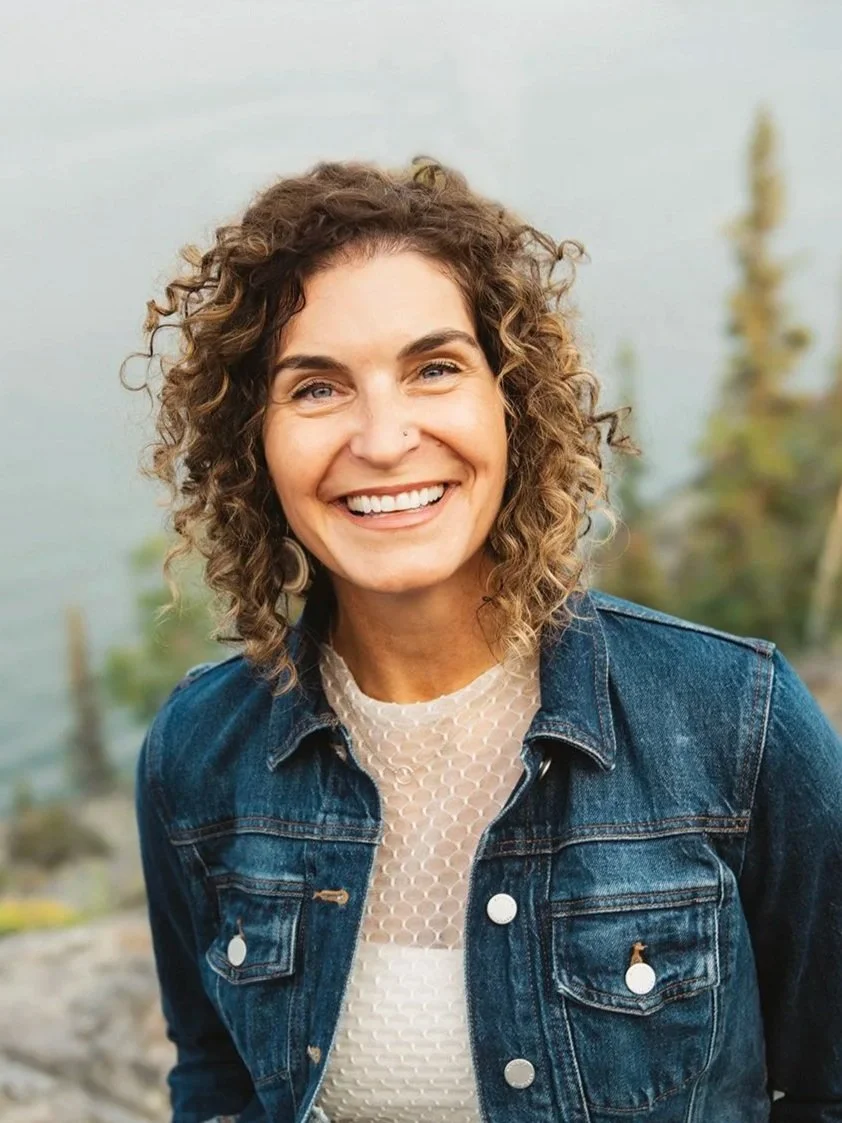 A woman with curly brown hair smiling outdoors near a body of water and trees, wearing a denim jacket and white top.