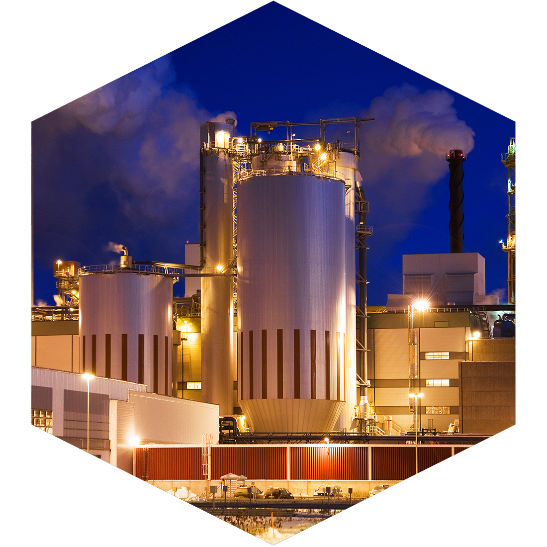 Night view of a large industrial factory with tall metallic storage tanks and chimneys emitting smoke, illuminated by bright lights.
