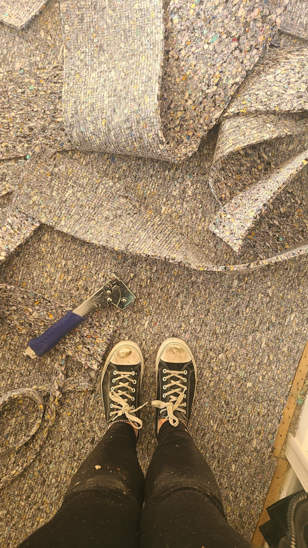 A pair of worn black and white sneakers on a multicolored, speckled carpeted floor, with a staple gun and a roll of fabric or carpet nearby, indicating an ongoing flooring or upholstery project.
