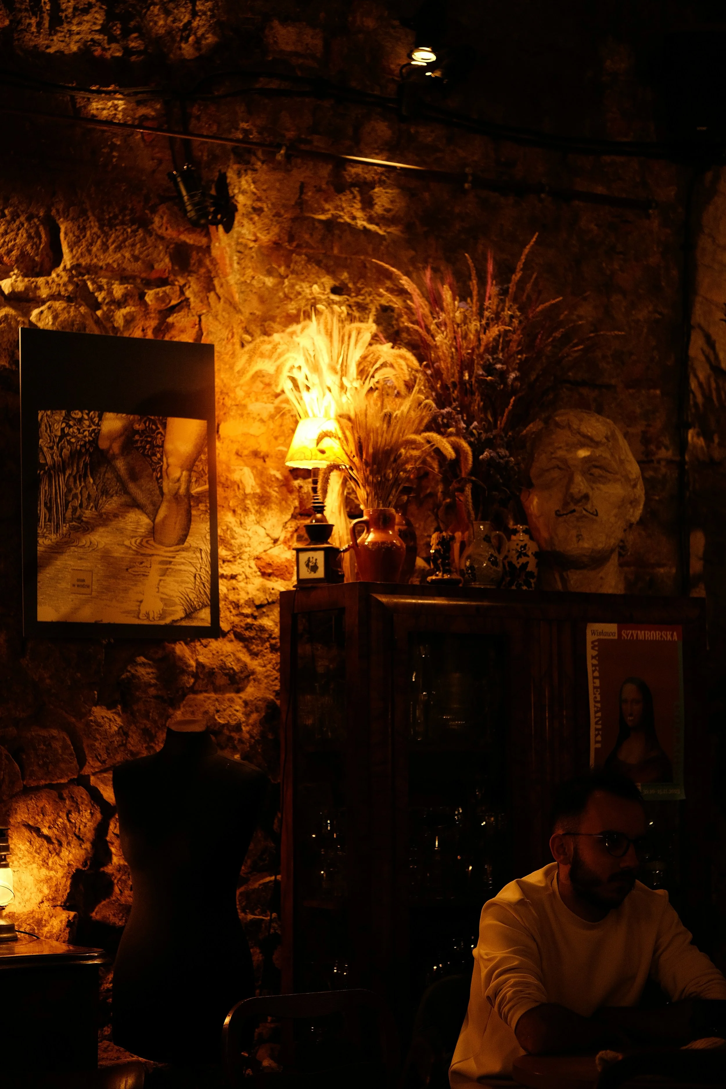 Interior of a dimly lit room with a stone wall, decorated with a framed picture, a sculpture of a face, and vases with dried plants, with a man sitting at a table.