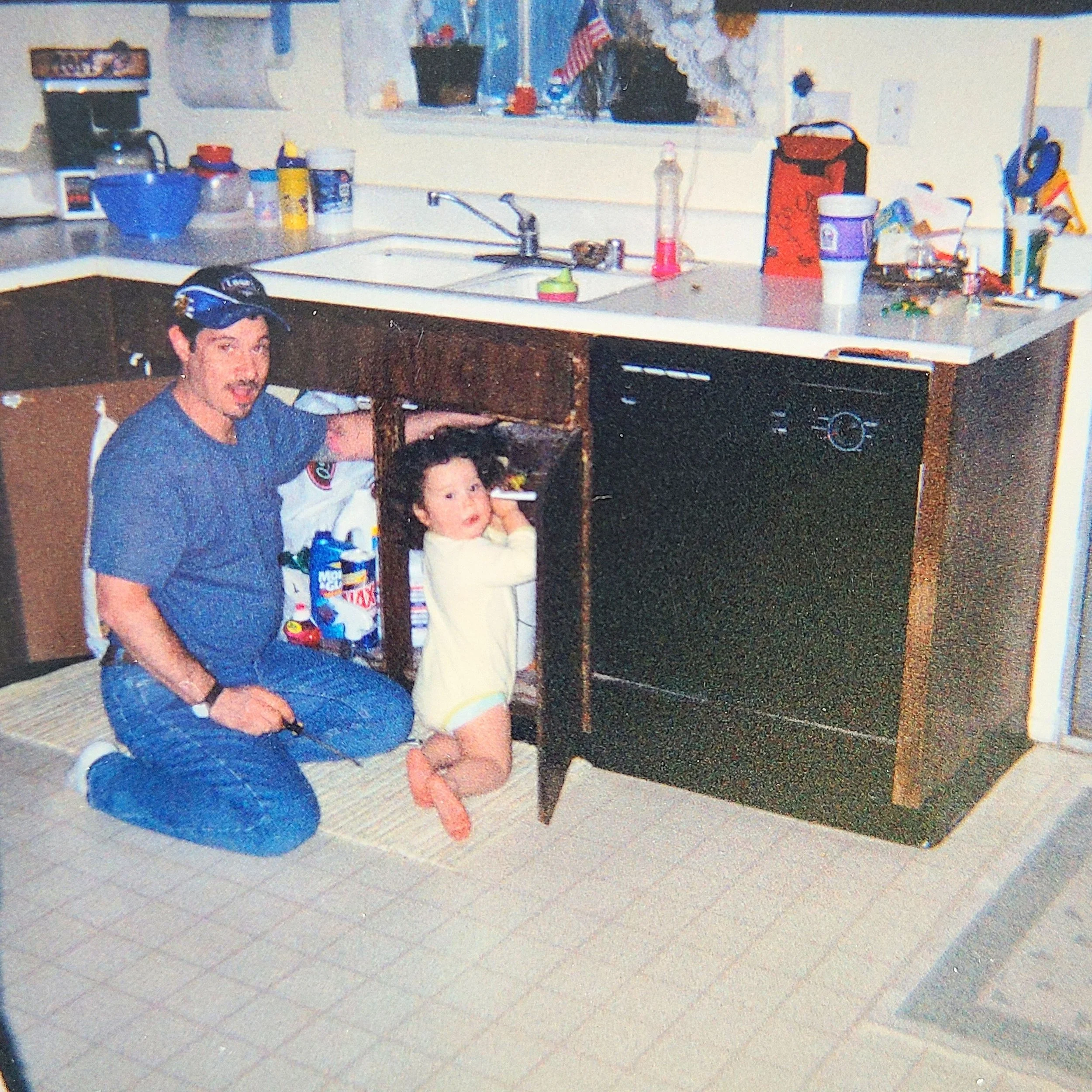 A man kneeling on the floor with a young girl next to him, in a kitchen with open cabinet under the sink. The man is wearing a dark baseball cap and blue shirt, the girl is wearing a yellow dress. The kitchen has various items on the countertop, and a window above the sink.