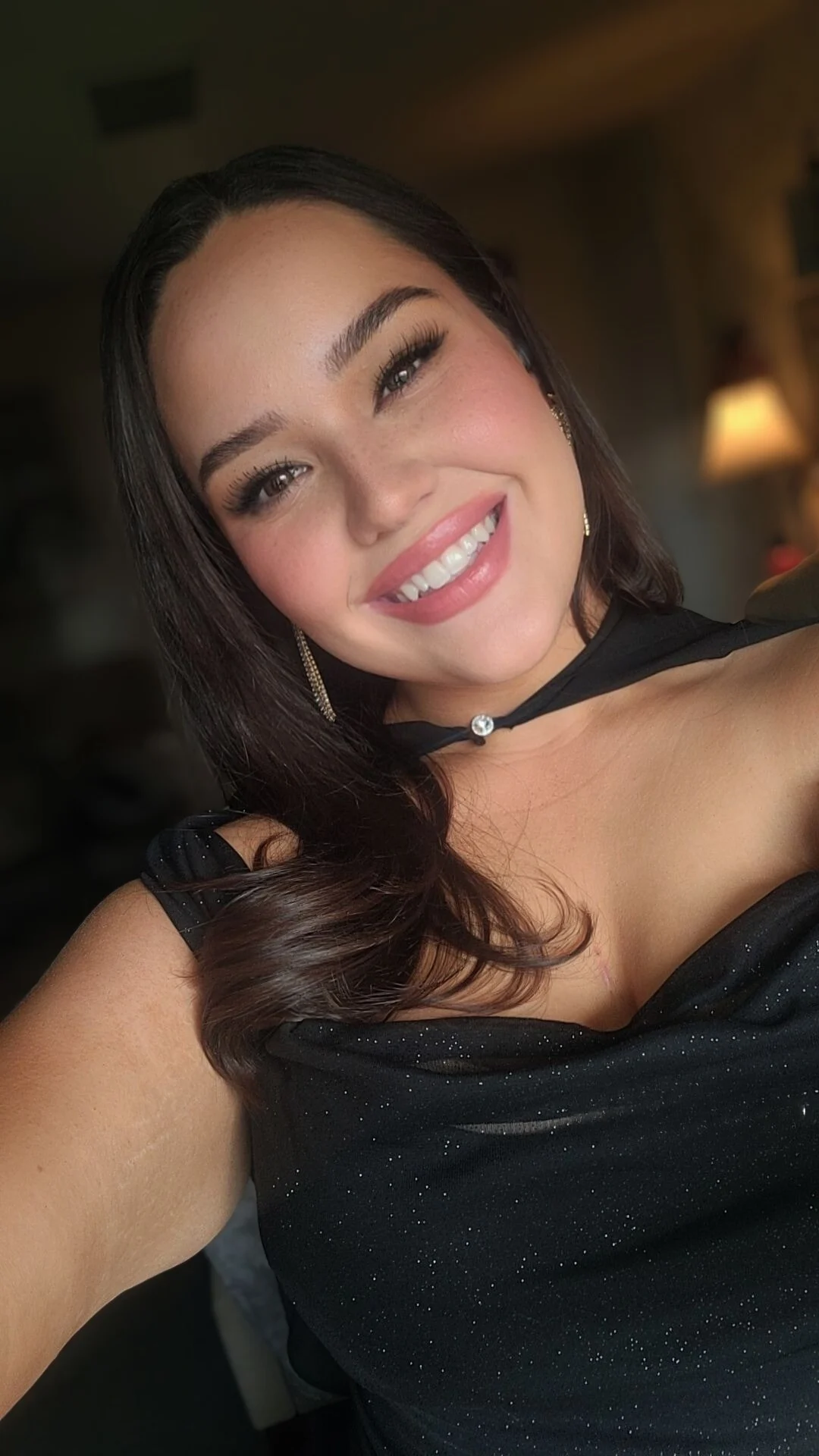 A young woman with dark hair and makeup smiling at the camera, wearing a black dress with sparkles and gold earrings, indoors with warm lighting in the background.