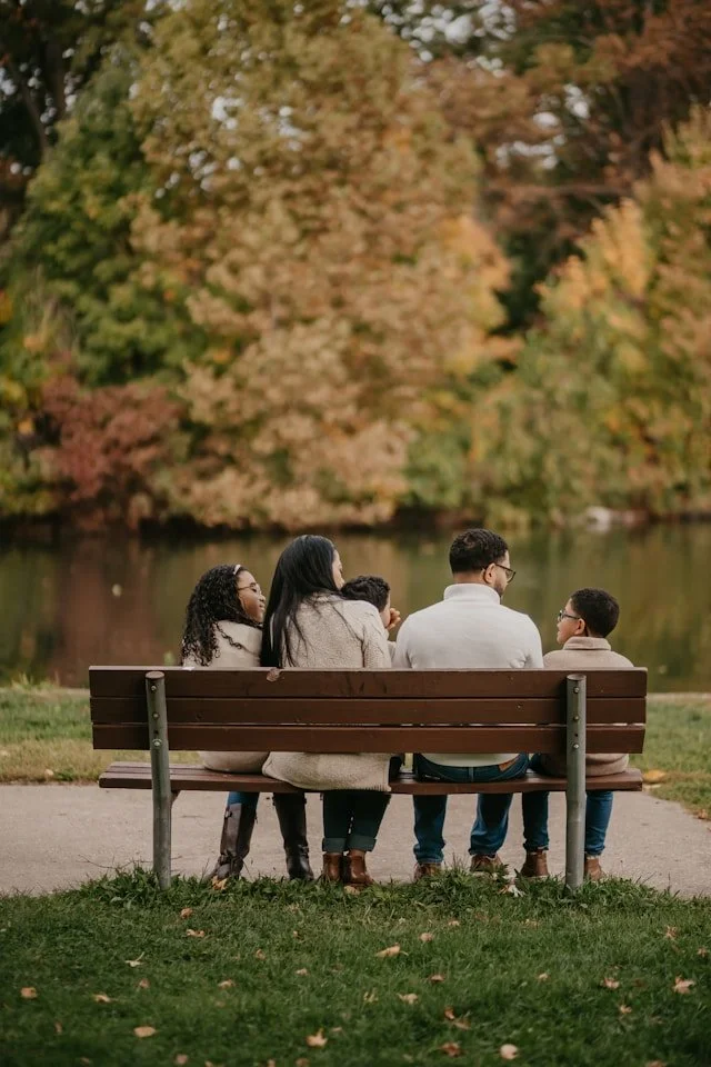 Family sitting on a park bench by a lake during autumn with colorful fall foliage in the background.