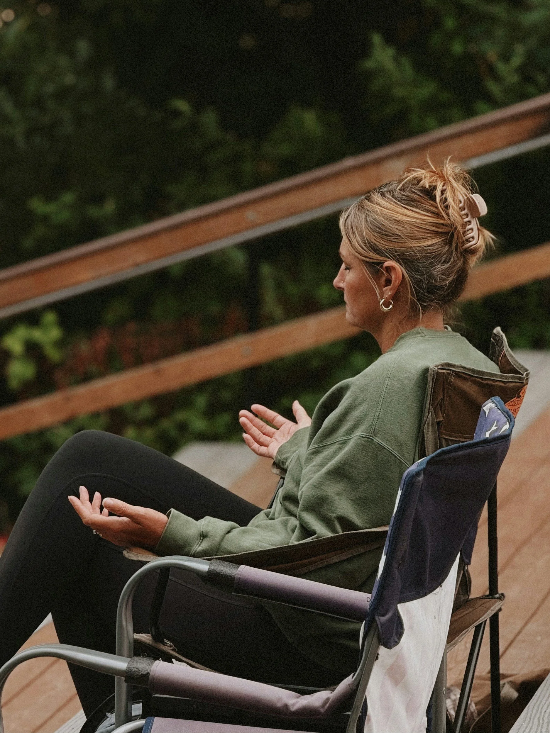 A woman seated in a lawn chair with her eyes closed, hands resting on her lap, and wearing a hair clip and hoop earrings. The background is blurred greenery.