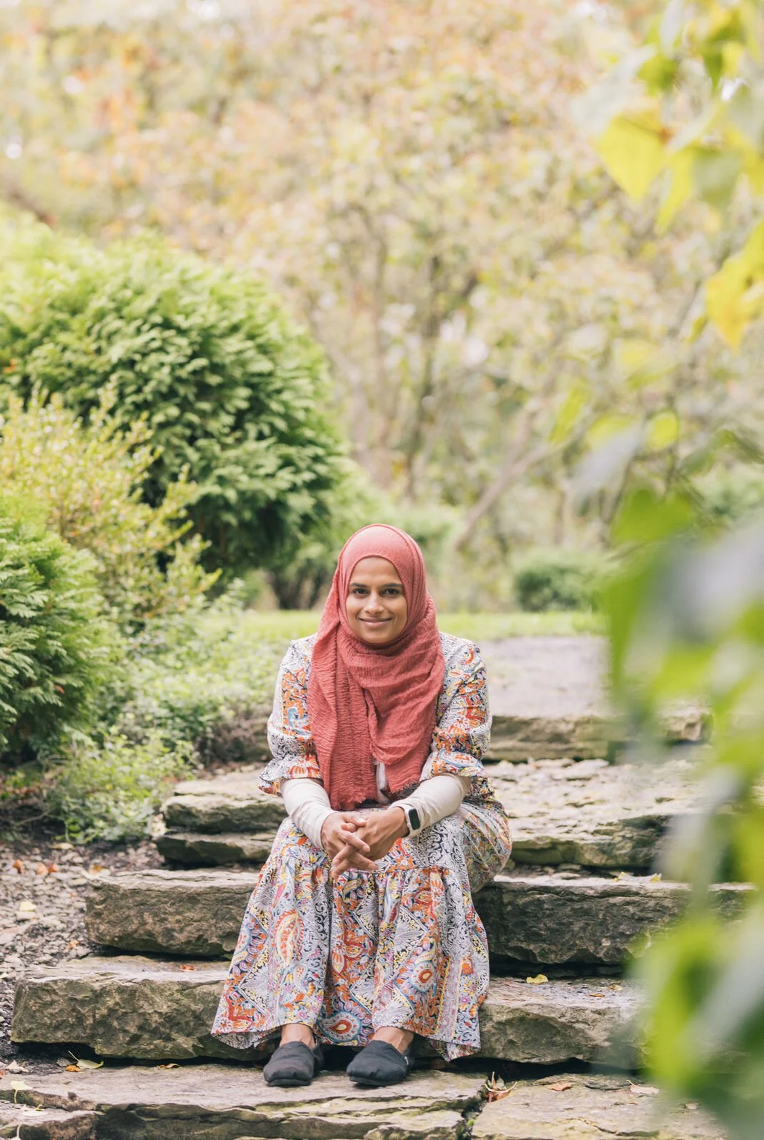 A woman wearing a pink headscarf, patterned dress, and black shoes sitting on stone steps outdoors in a green park.