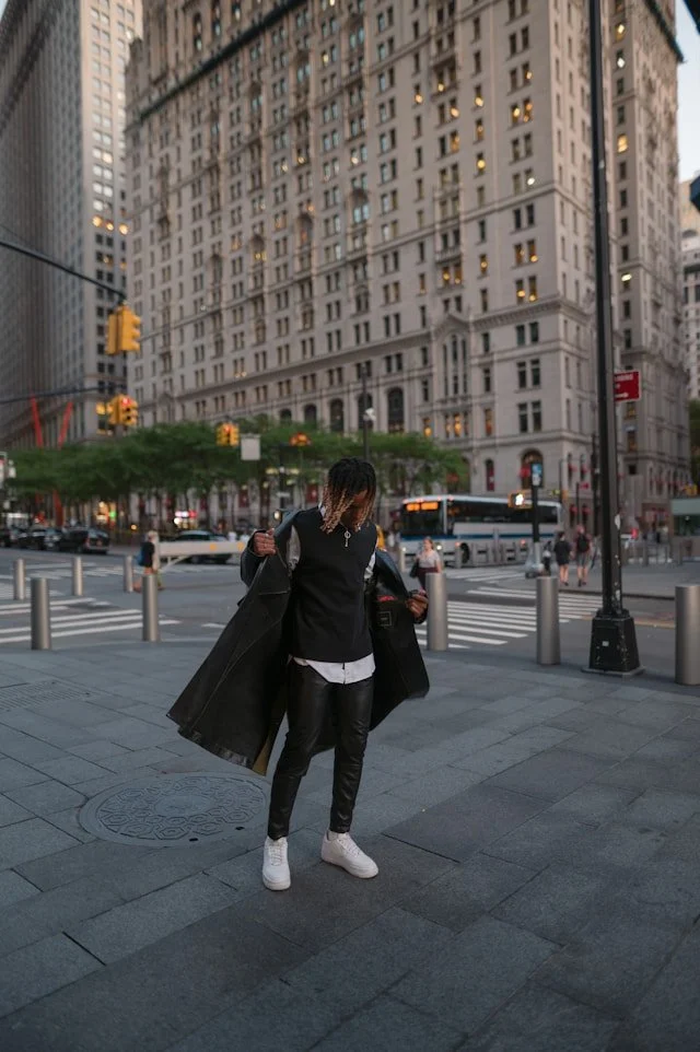 Person in black outfit and white sneakers standing on city sidewalk at dusk, with tall buildings and traffic in the background.