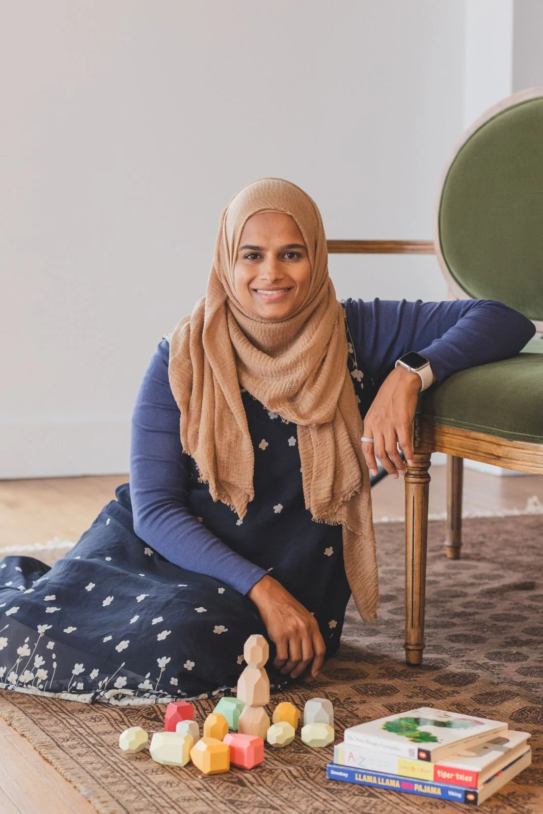A woman wearing a beige hijab and a dark dress with white floral patterns, sitting on a patterned rug with children's wooden toys and books, smiling indoors.