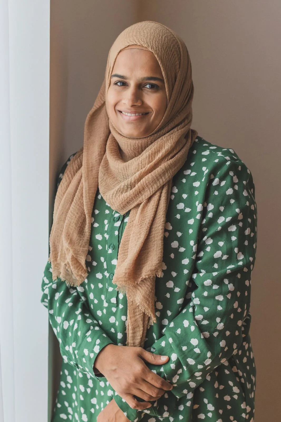 A woman wearing a beige hijab and a green dress with white dot patterns, smiling and standing by a wall.