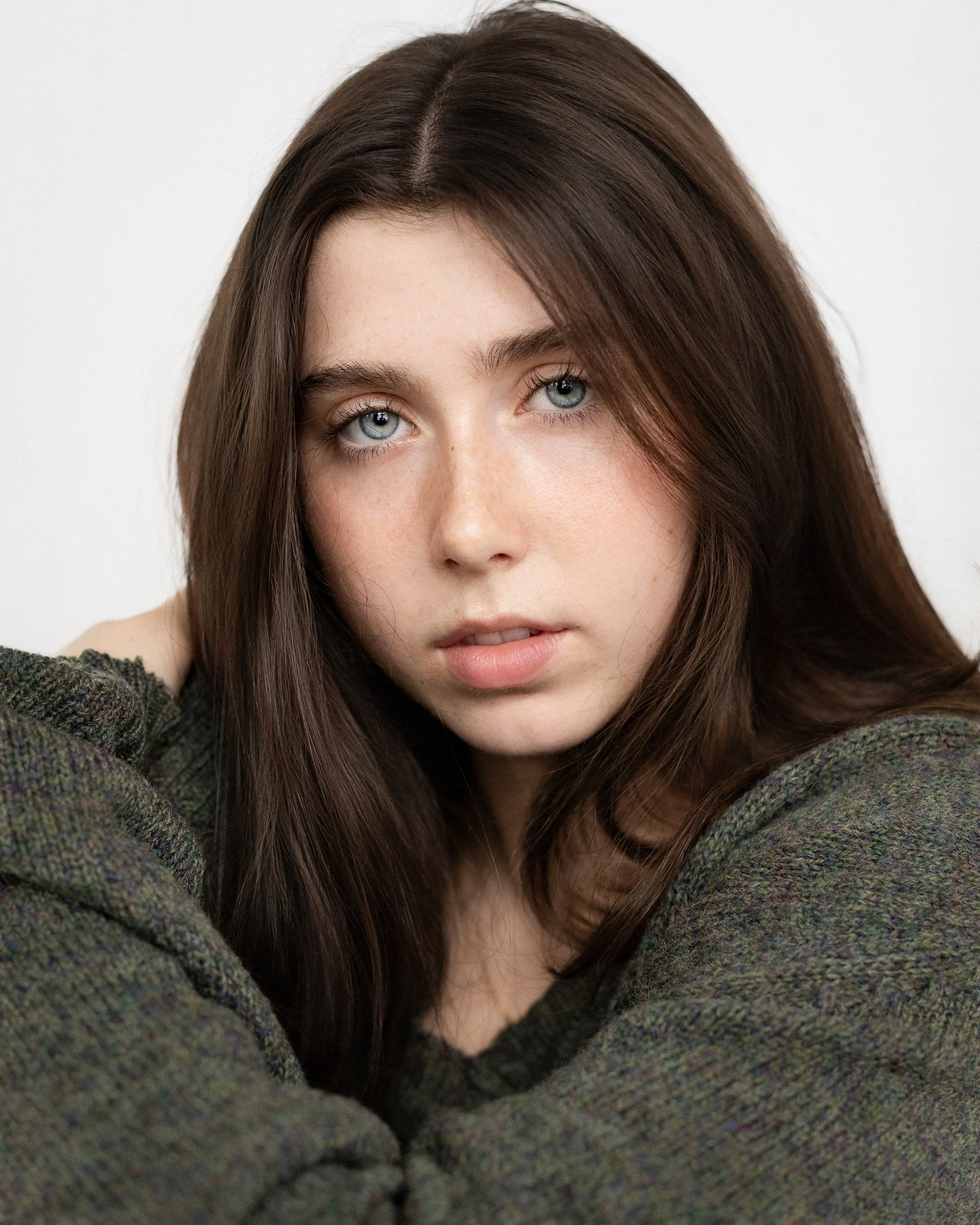 Close-up portrait of a young woman with blue eyes and long brown hair, wearing a dark green sweater, looking directly at the camera with a neutral expression.
