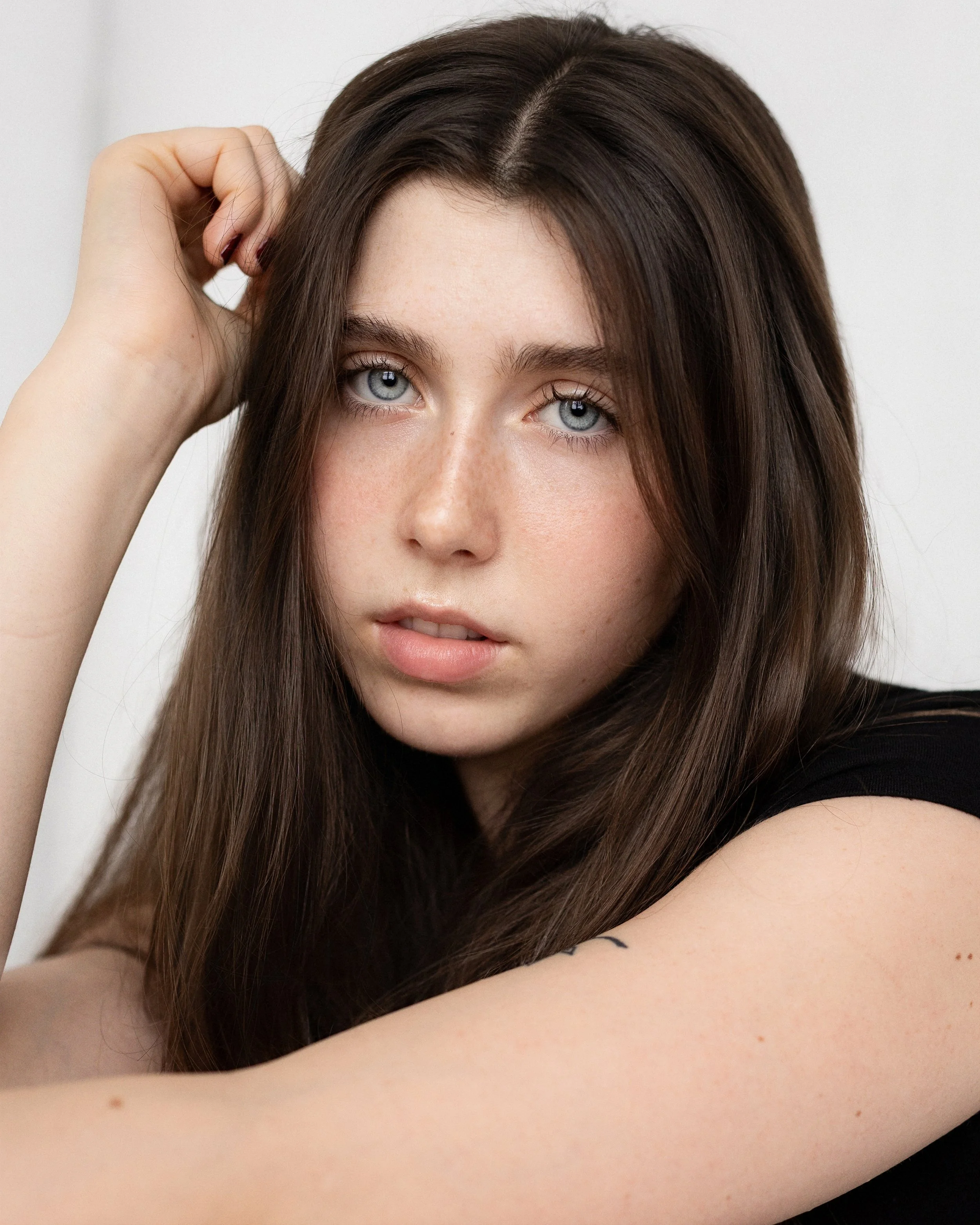 Close-up of a young woman with long brown hair and blue eyes, looking at the camera with her hand near her head.