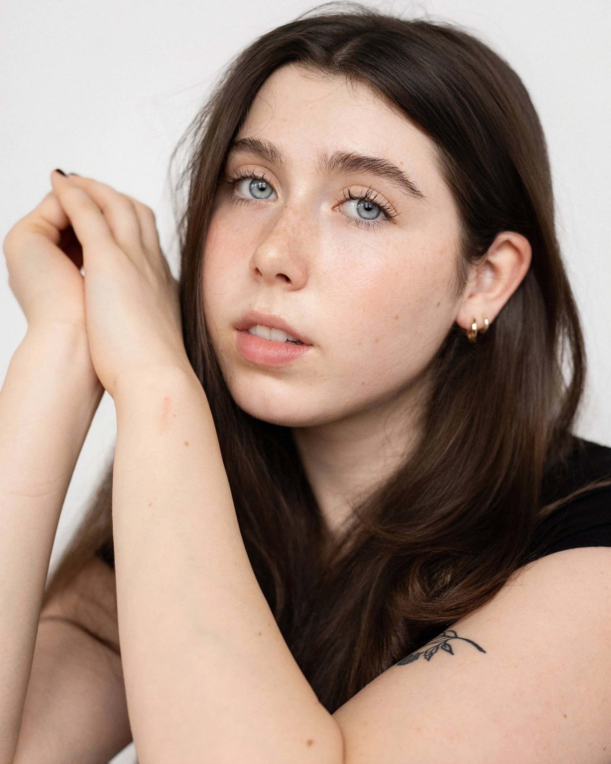 Close-up of a young woman with blue eyes and long brown hair, resting her chin on her hands, against a plain white background.