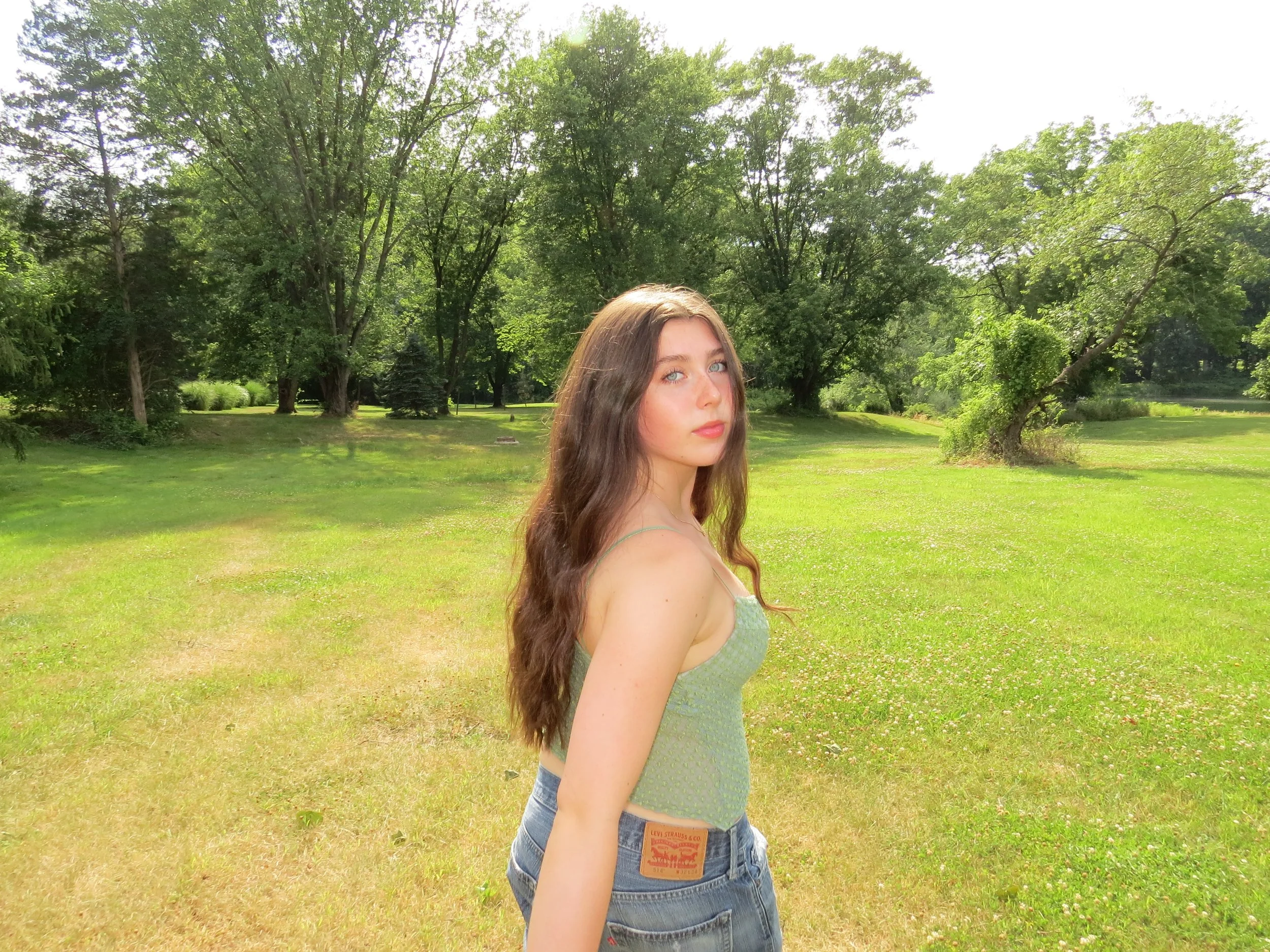 A young woman with long wavy brown hair and blue eyes standing in a green park with trees and grass, looking at the camera.