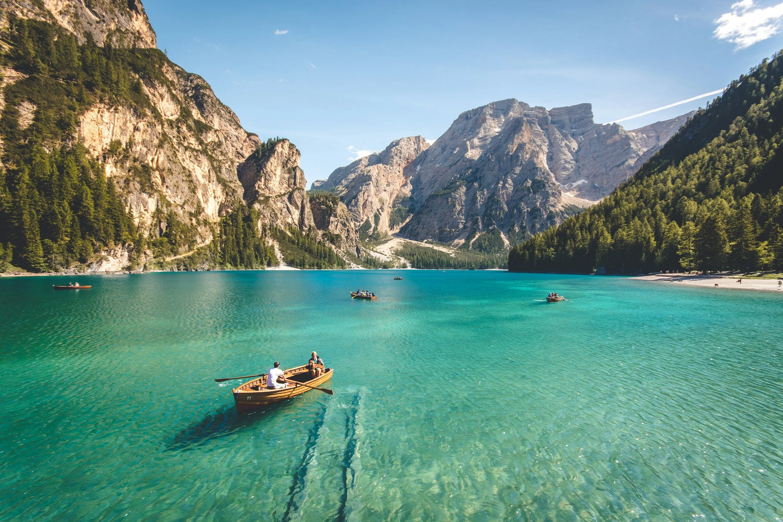 Scenic view of a turquoise lake surrounded by mountains and evergreen trees, with several boats floating on the water and a small sandy beach on the right side.