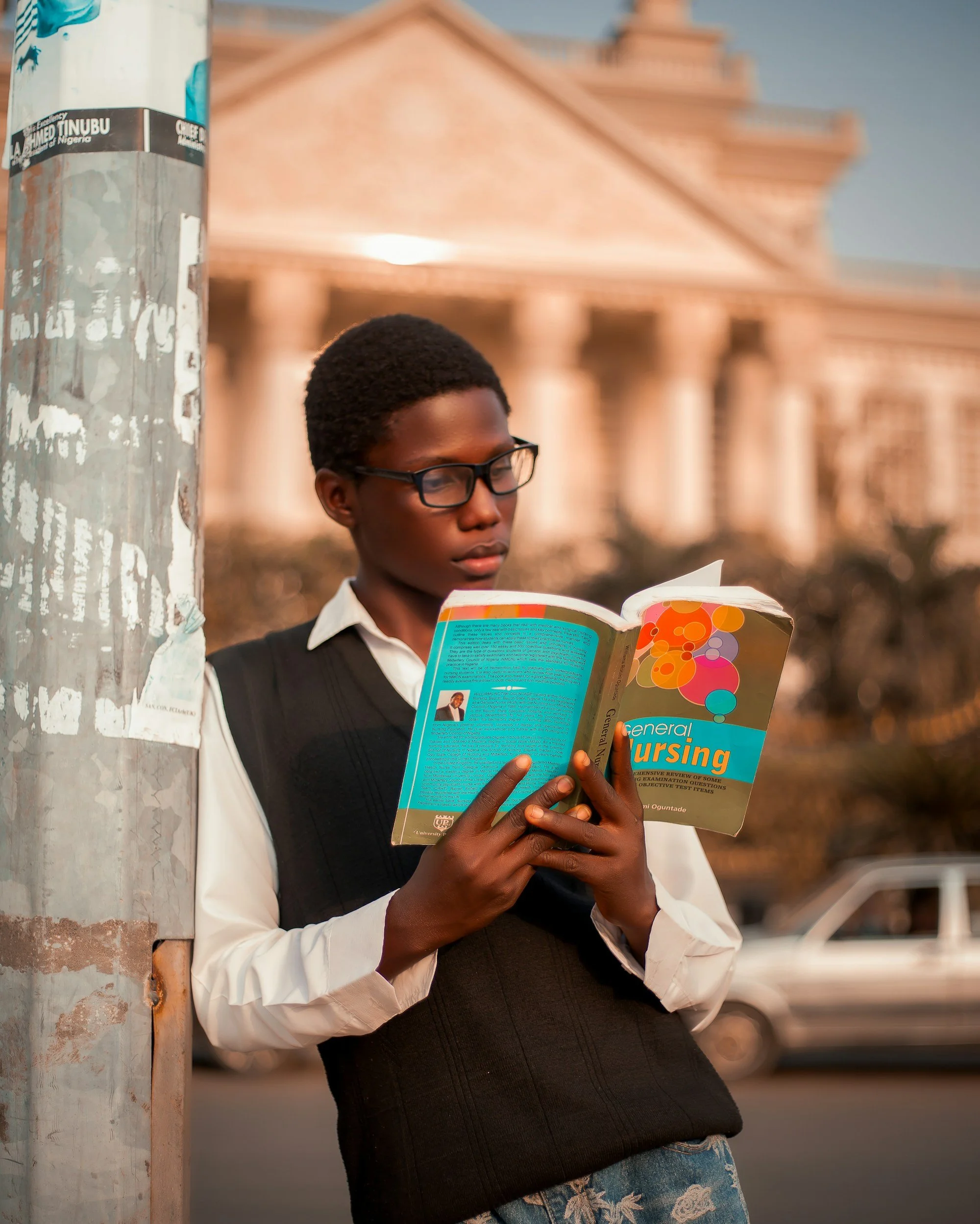 A young person with glasses reading a book titled 'General Nursing' while leaning against a metal pole on a city street, with a classical building in the background.