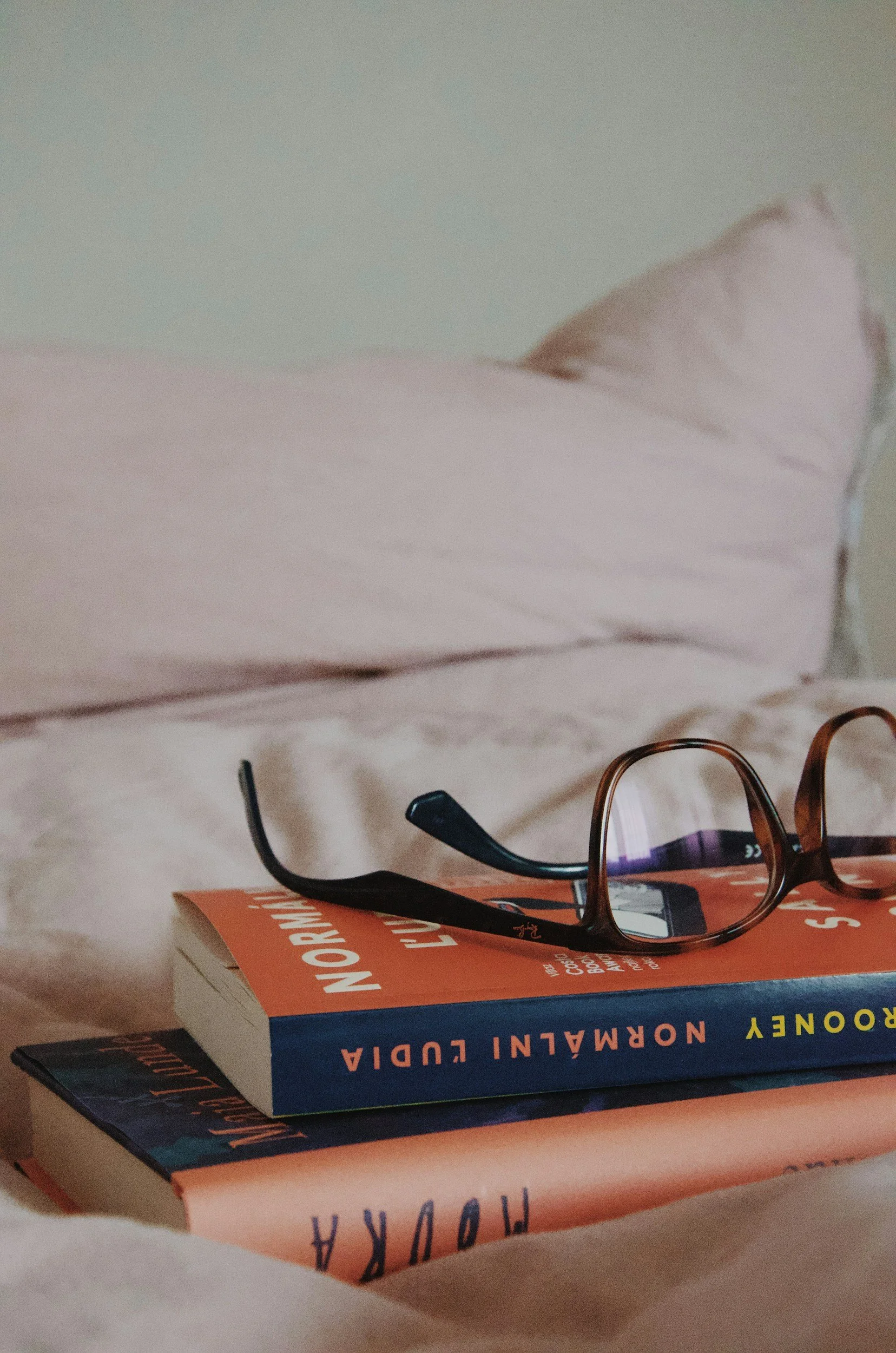 A pair of eyeglasses resting on top of a stack of books on a bed with a pink pillow in the background.