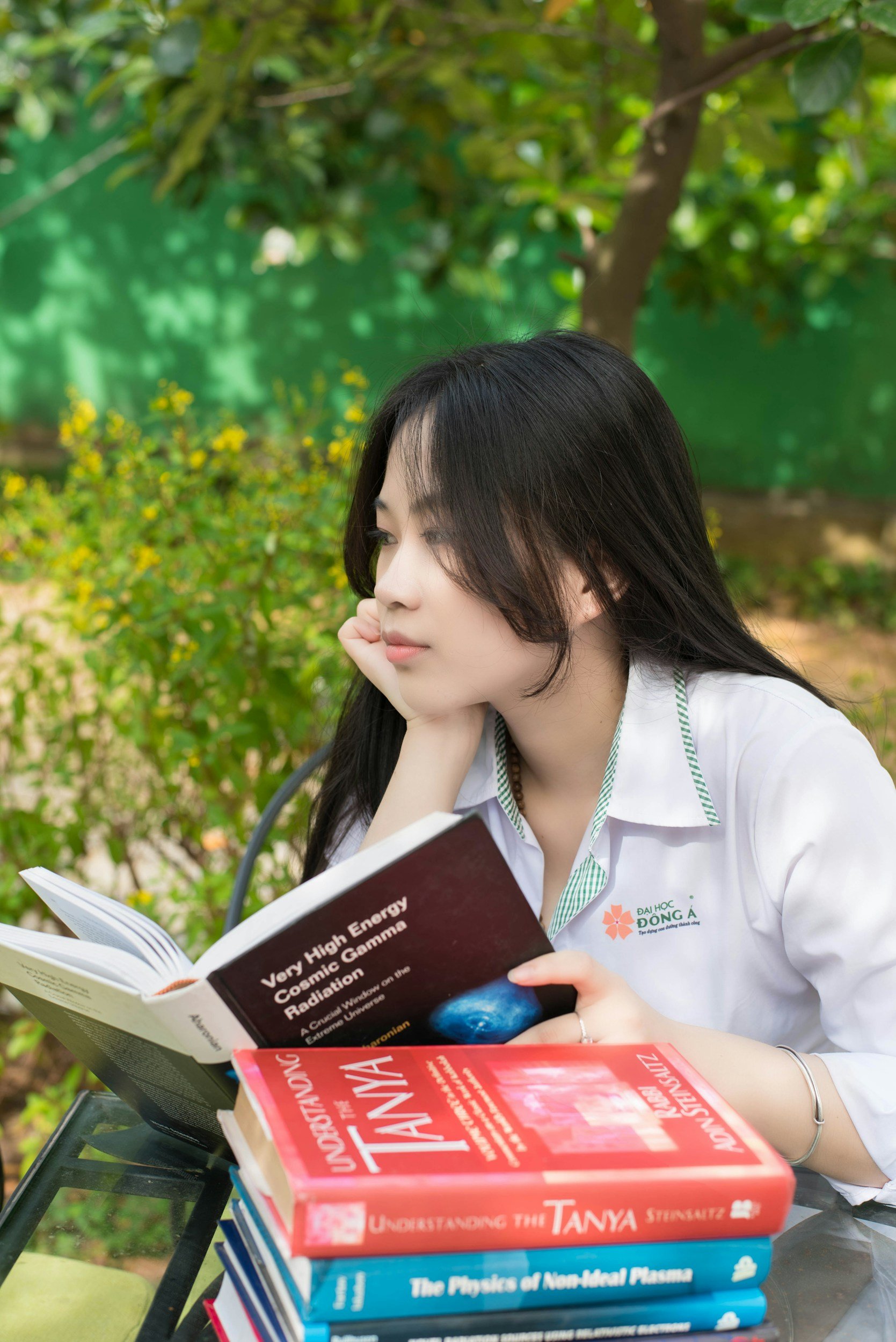 A young woman with long black hair reading a book outdoors, sitting at a table with a stack of books in front of her, surrounded by greenery.