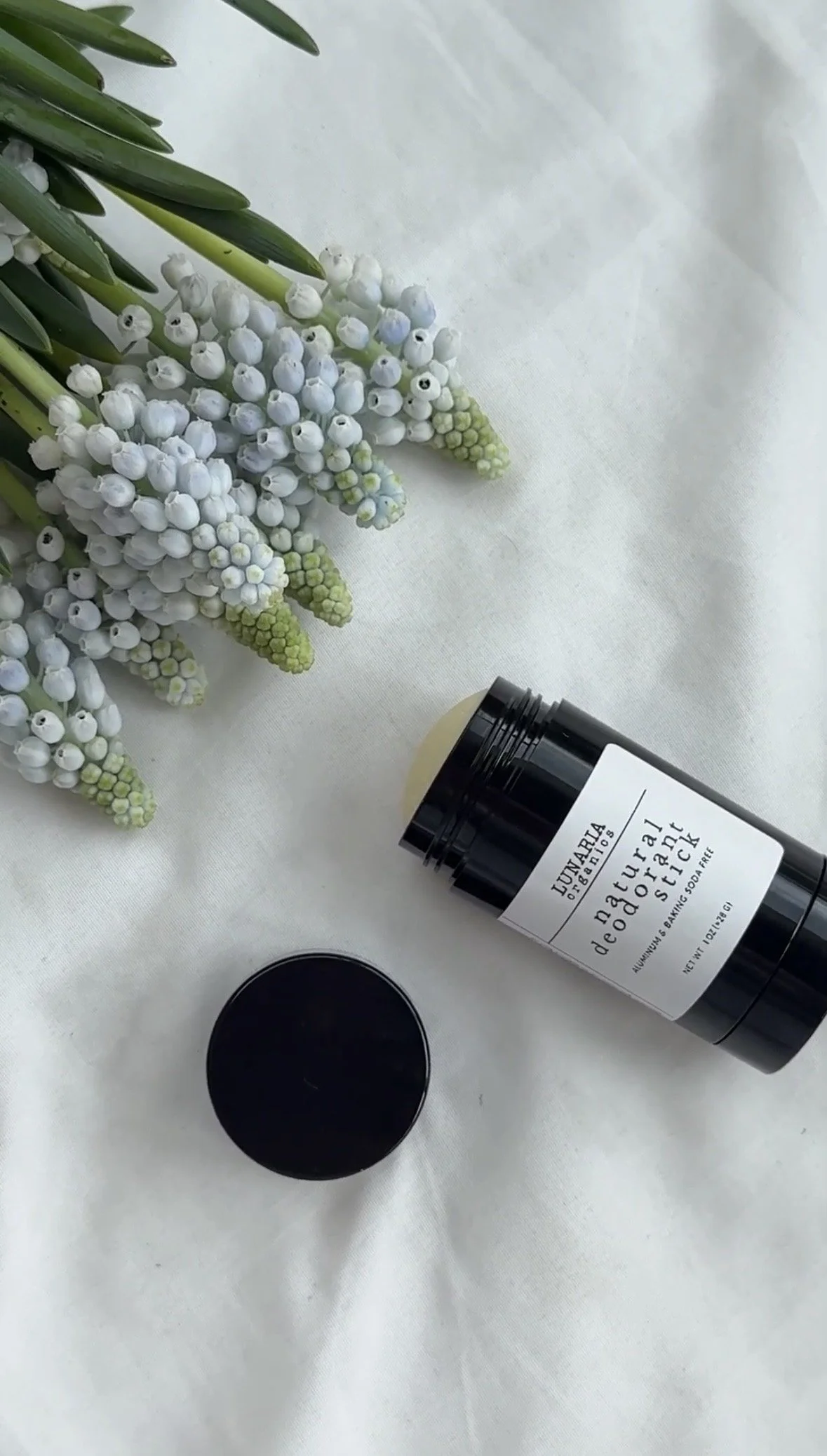 A linen or fabric surface with a bunch of white and light purple grape hyacinth flowers and a black container labeled 'Humble Brands Natural Deodorant' with its lid placed nearby.
