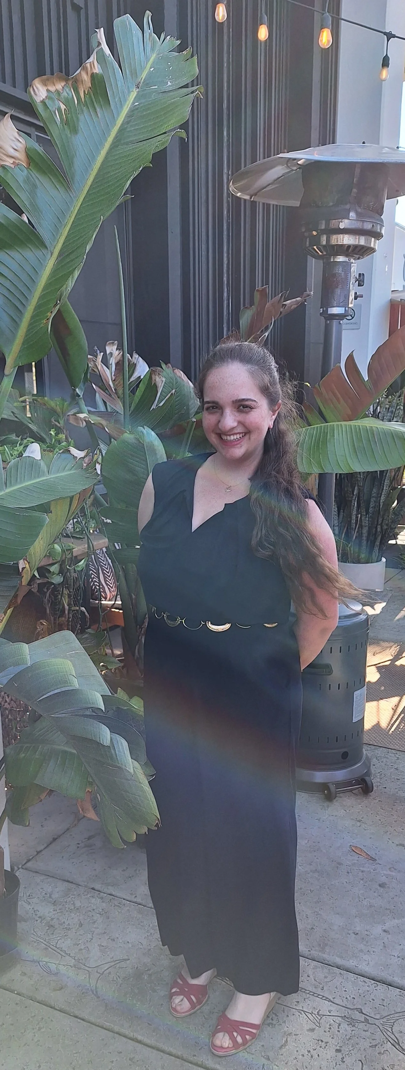 A smiling woman with long, dark, curly hair in a black dress standing outdoors near tropical plants and a patio heater, with string lights overhead.