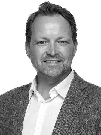 Schwarz-weiß-Foto eines Mannes mit braunen Haaren, Bart, weißem Hemd und grauem Blazer, lächelnd.